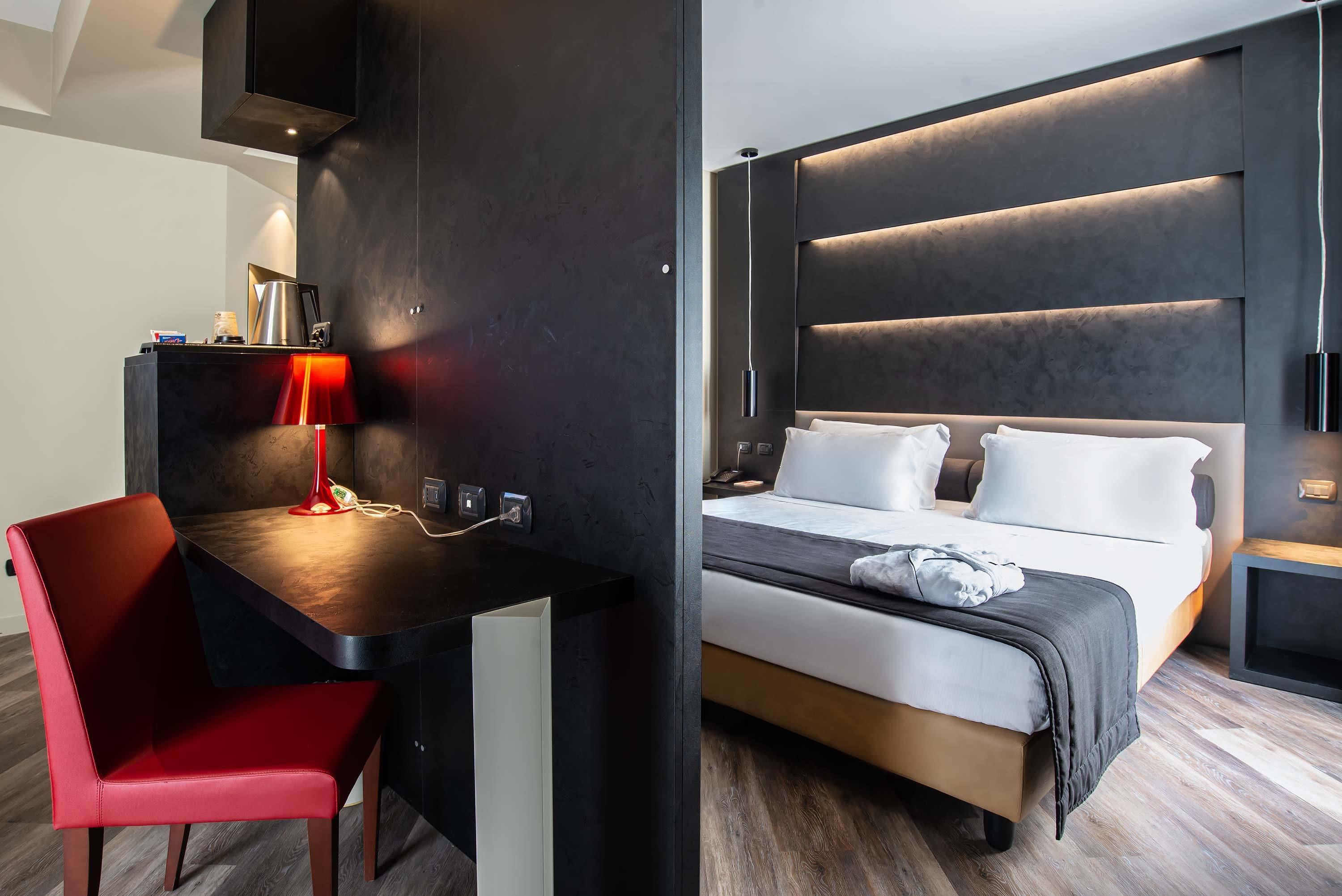 A modern hotel room with a bed, bedside table, red chair, and a desk with a lamp.
