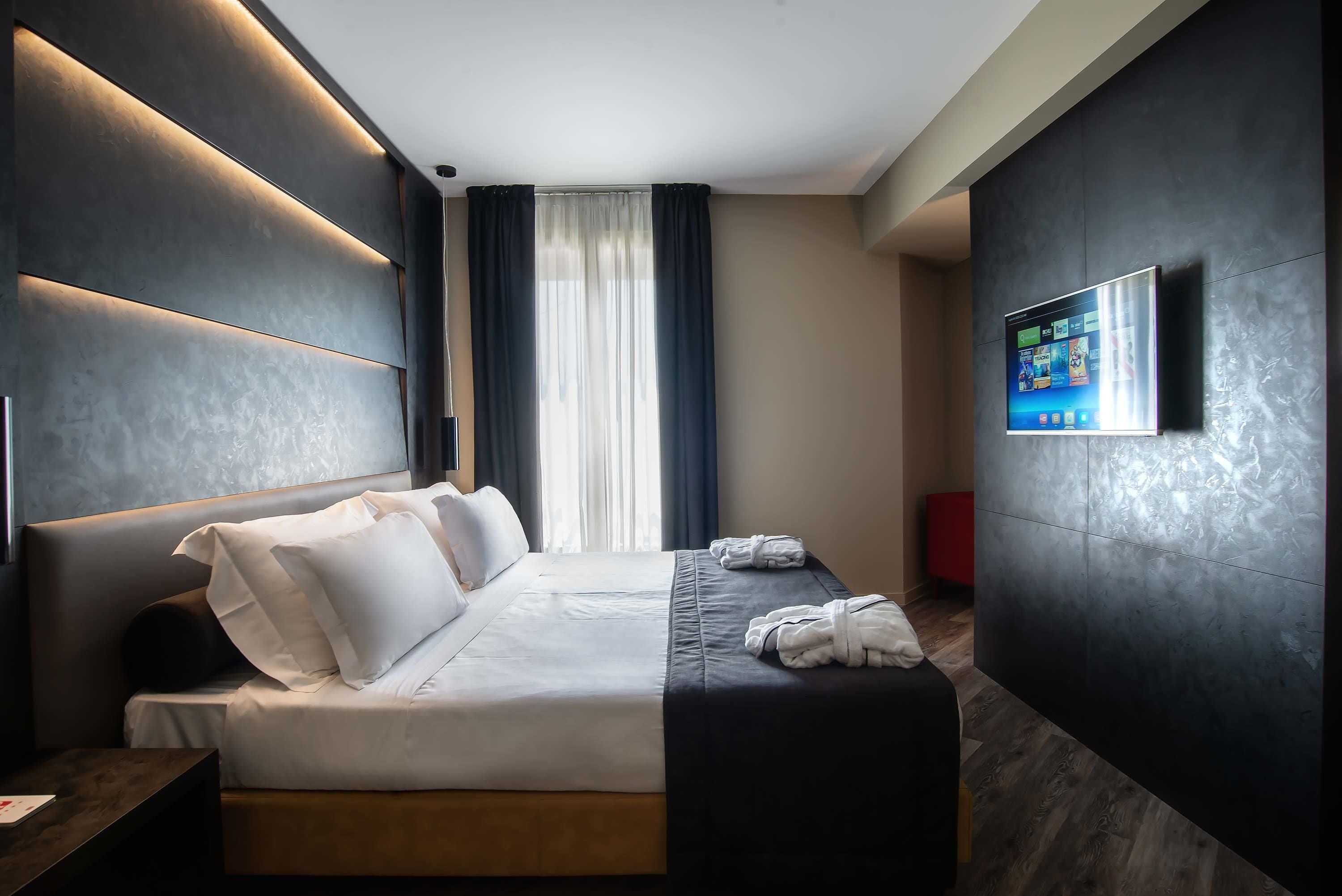 A modern hotel room with a large bed, a flat-screen TV mounted on the wall, and a view of the outdoors through the window.