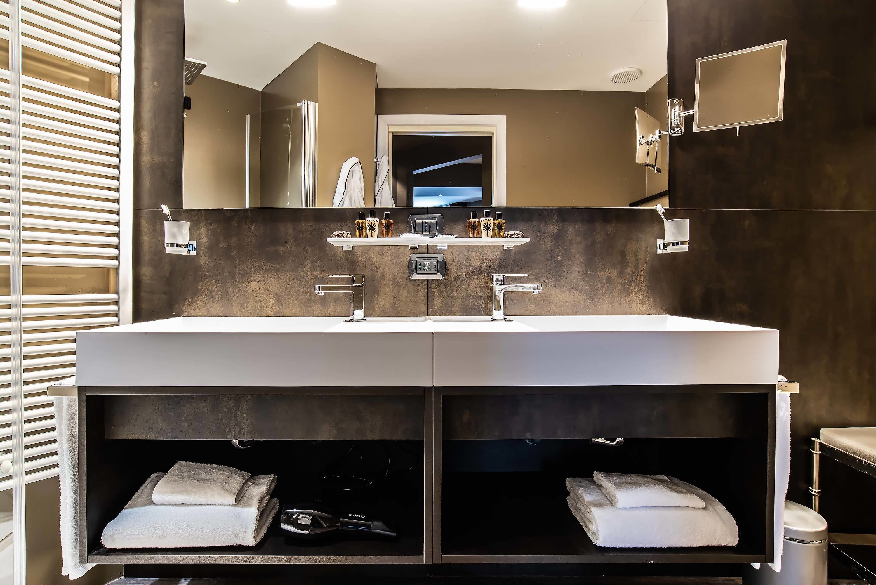 A modern bathroom with a large vanity, two sinks, and a mirror. There are towel racks and a shelf with toiletries.