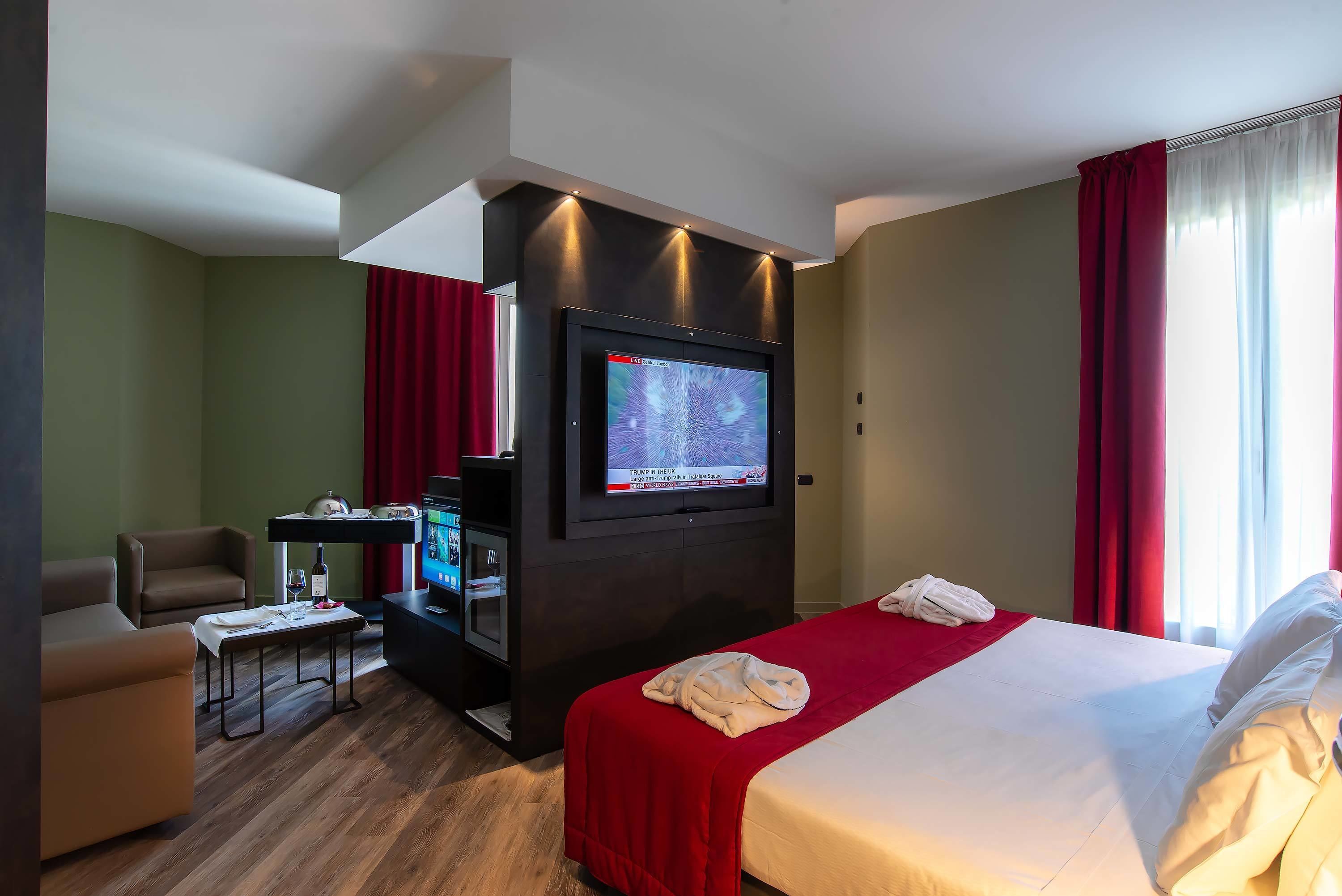 A hotel room with a bed, a TV mounted on the wall, a seating area with a table, and a red curtain.