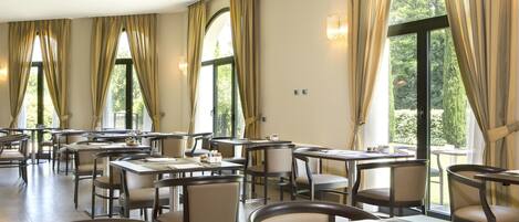 Comedor con mesas y sillas, ventanas amplias con cortinas y vistas a zonas verdes.