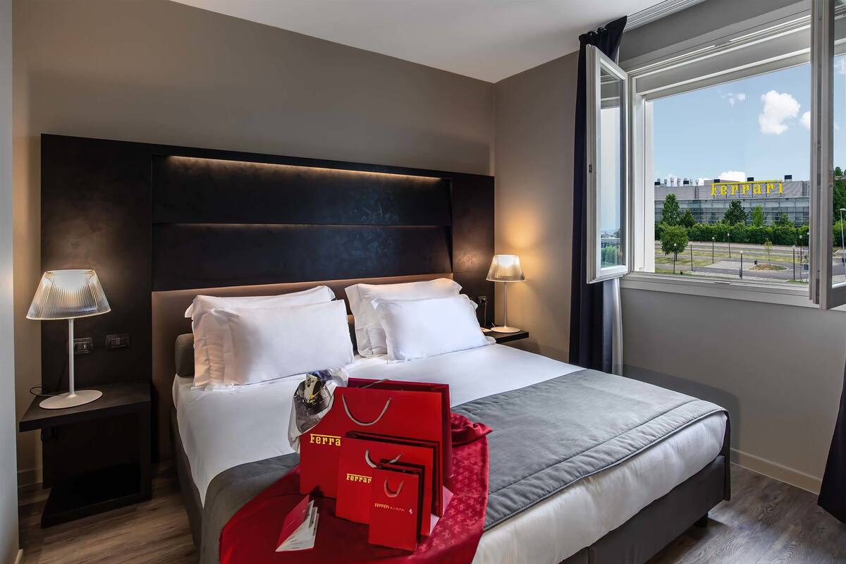 A modern hotel room with a large bed, two bedside tables with lamps, a window with a view of a building, and red shopping bags on the bed.