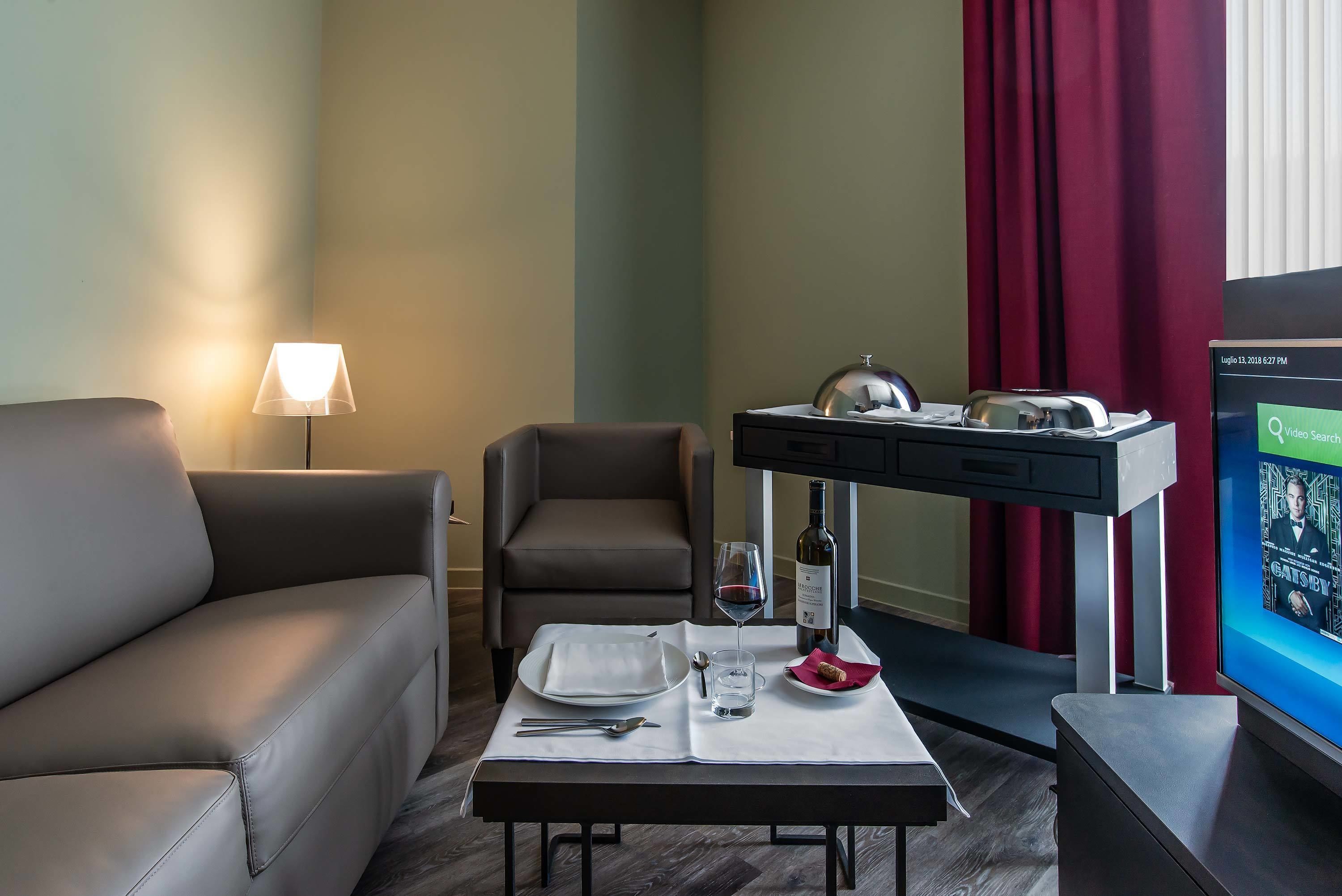 Ein Hotelzimmer mit Sofa, einem Esstisch gedeckt mit Tellern, Gläsern und einer Flasche, einem Fernseher und einem Schreibtisch mit Lampe.