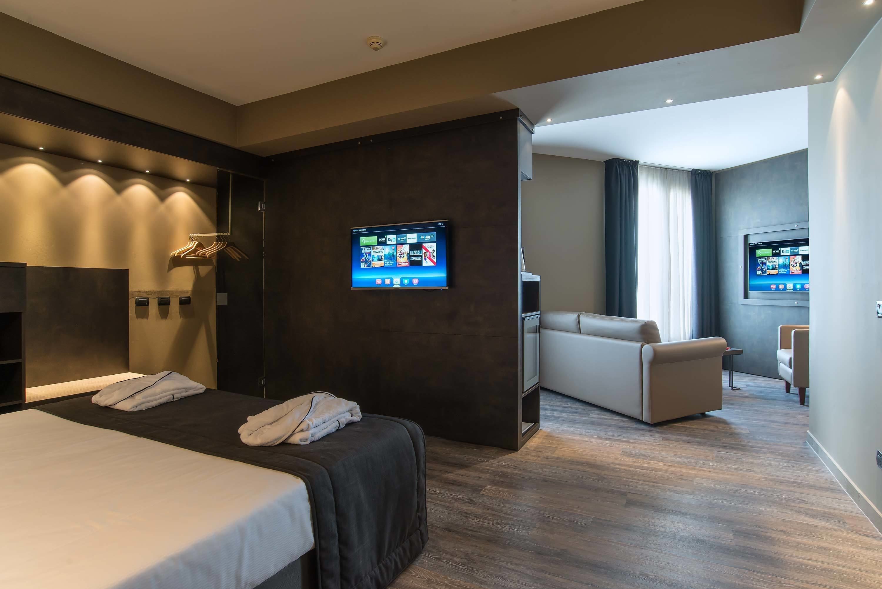 A modern hotel room with a large bed, a seating area, and two flat-screen TVs mounted on the wall.