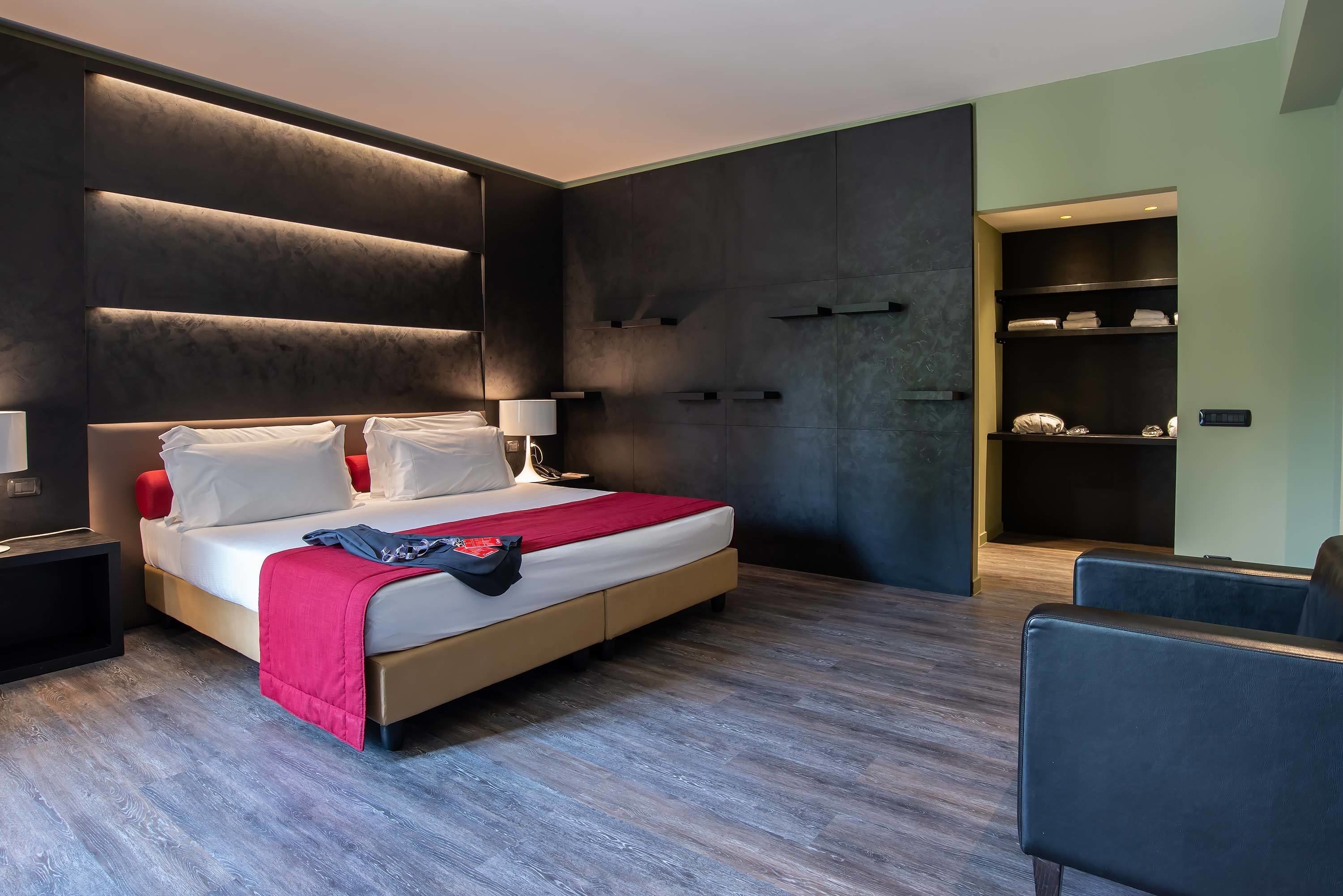 A modern hotel room with a large bed, bedside tables, a nightstand, a built-in wardrobe, and a seating area.