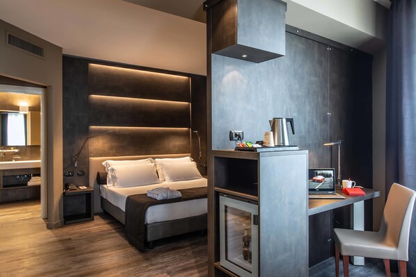A modern hotel room with a bed, a desk with a laptop, a chair, and a minibar.