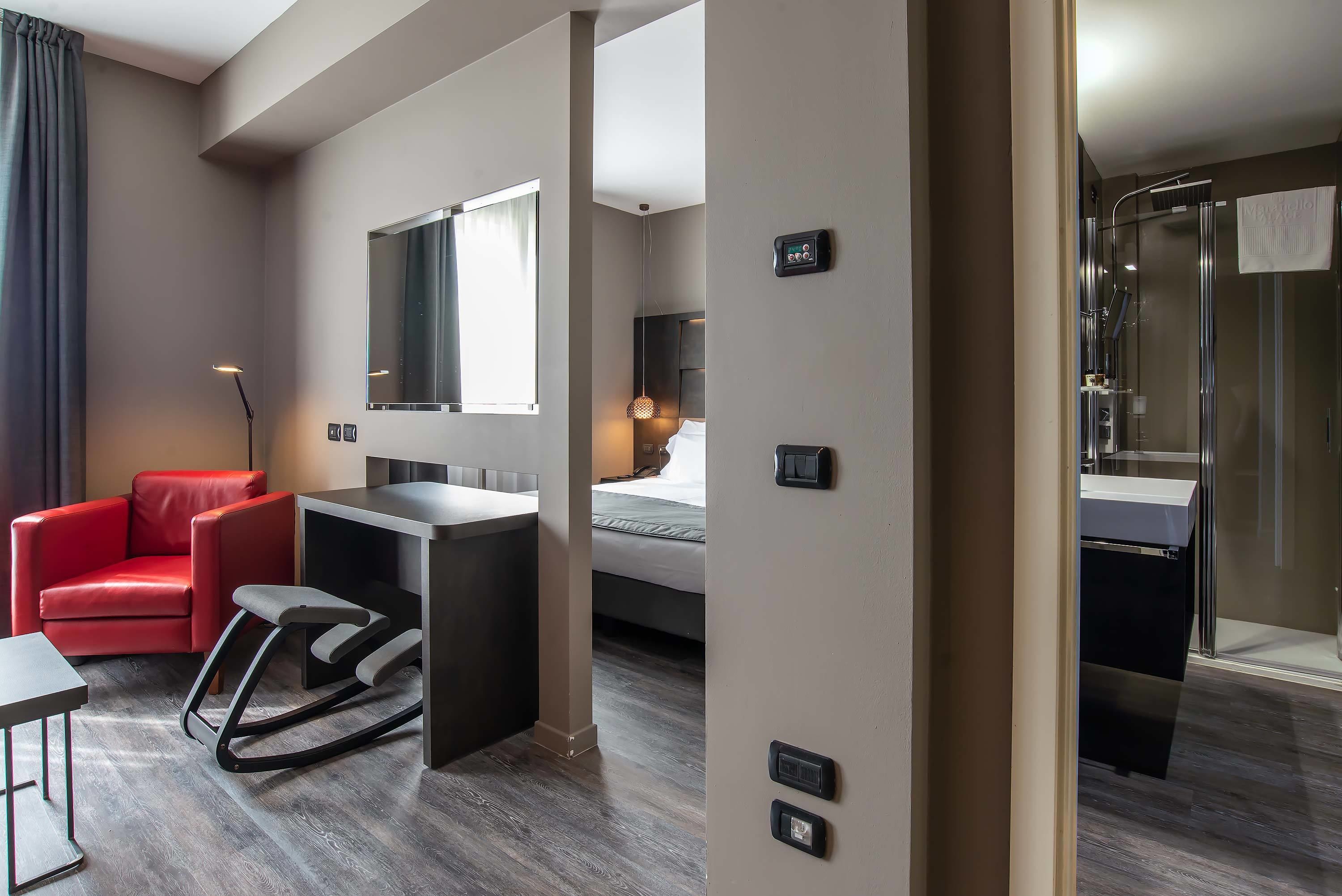 Una habitación de hotel moderna con una silla roja, un escritorio pequeño, una cama con cabecera, un espejo, zona de ducha y un panel de control en la pared.