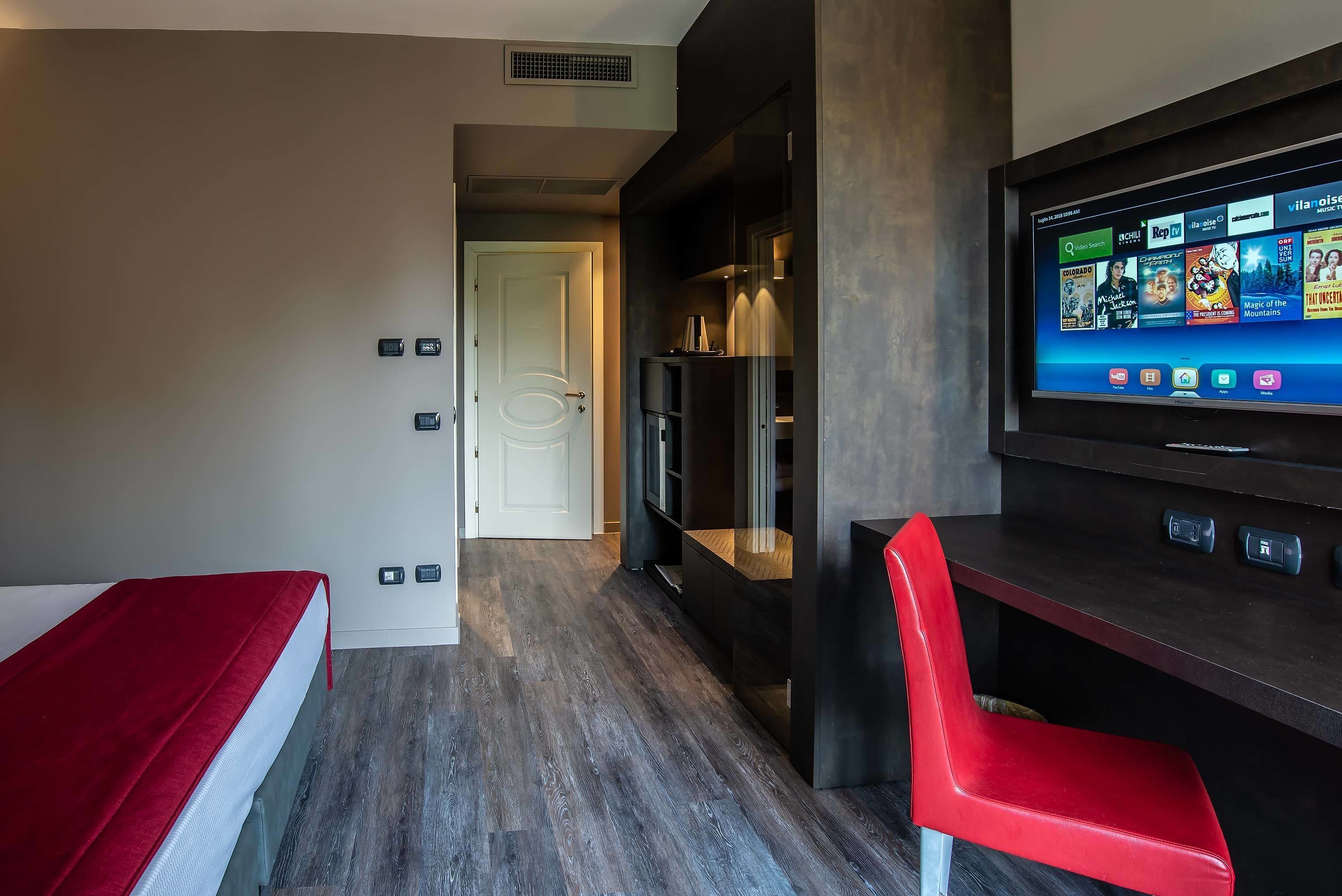 A hotel room with a bed, a TV, a red chair, and a minibar.