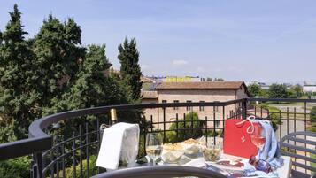 A balcony with a table set for a meal, overlooking a building with a red roof and greenery in the background.