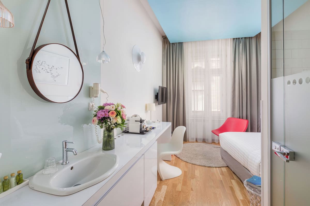 Moderni hotellihuone, jossa on valkoinen pesuallas, peili, punainen tuoli, sänky ja suuri ikkuna, jossa on verhot.