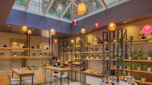 Un intérieur moderne avec un plafond haut et une verrière, comprenant un coin repas, un espace salon et des étagères ornées de plantes.