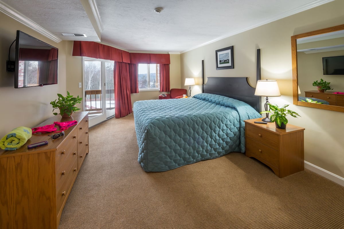 Une chambre d ’hôtel avec un grand lit, une commode en bois, un bureau avec une lampe, une plante en pot et une grande fenêtre avec des rideaux.