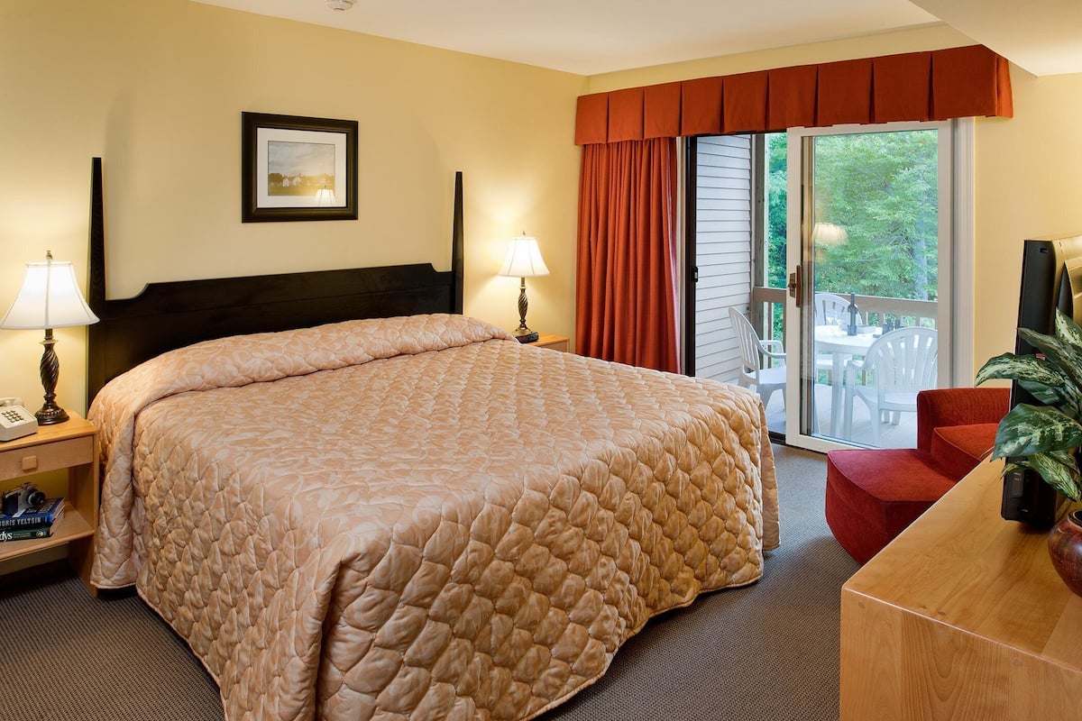 Une chambre d’hôtel avec un grand lit, deux lampes de chevet, un bureau avec une chaise, une plante et une porte-fenêtre coulissante donnant sur l’extérieur.