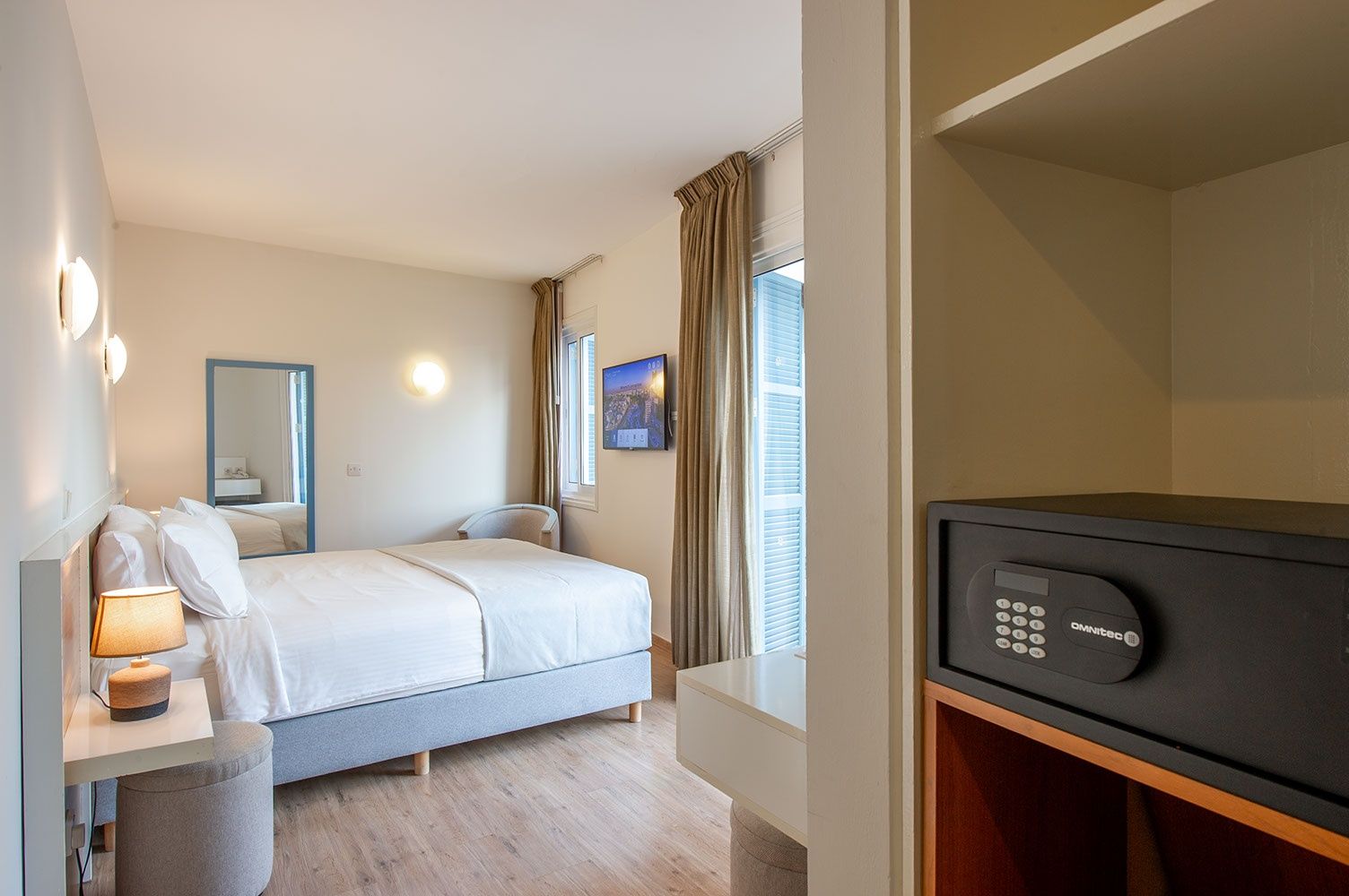A hotel room with a bed, bedside table with a lamp, a TV, and a wall-mounted control panel.
