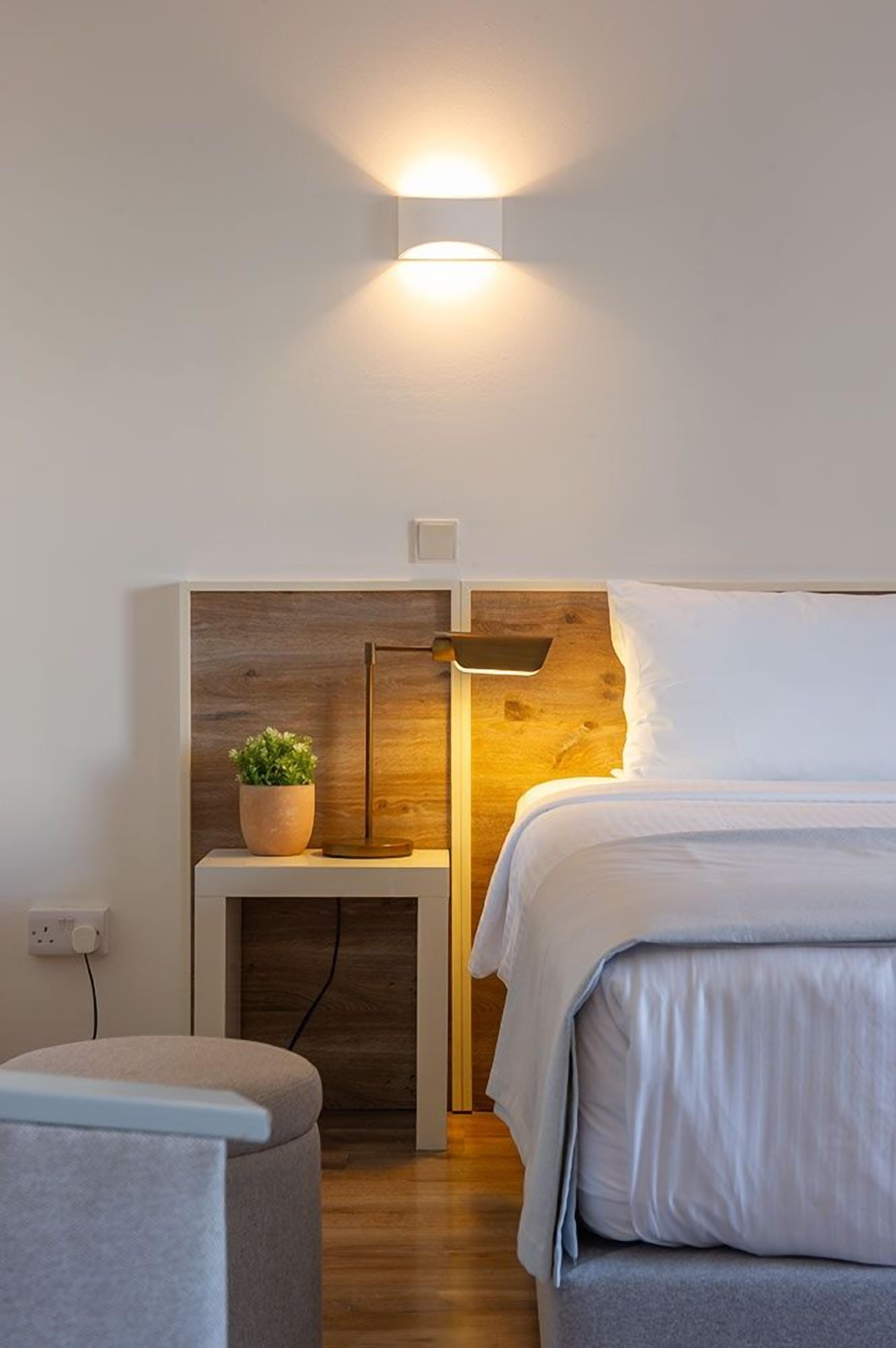 A bedroom with a bed, bedside table with a lamp, a small potted plant, and a wall-mounted light fixture.