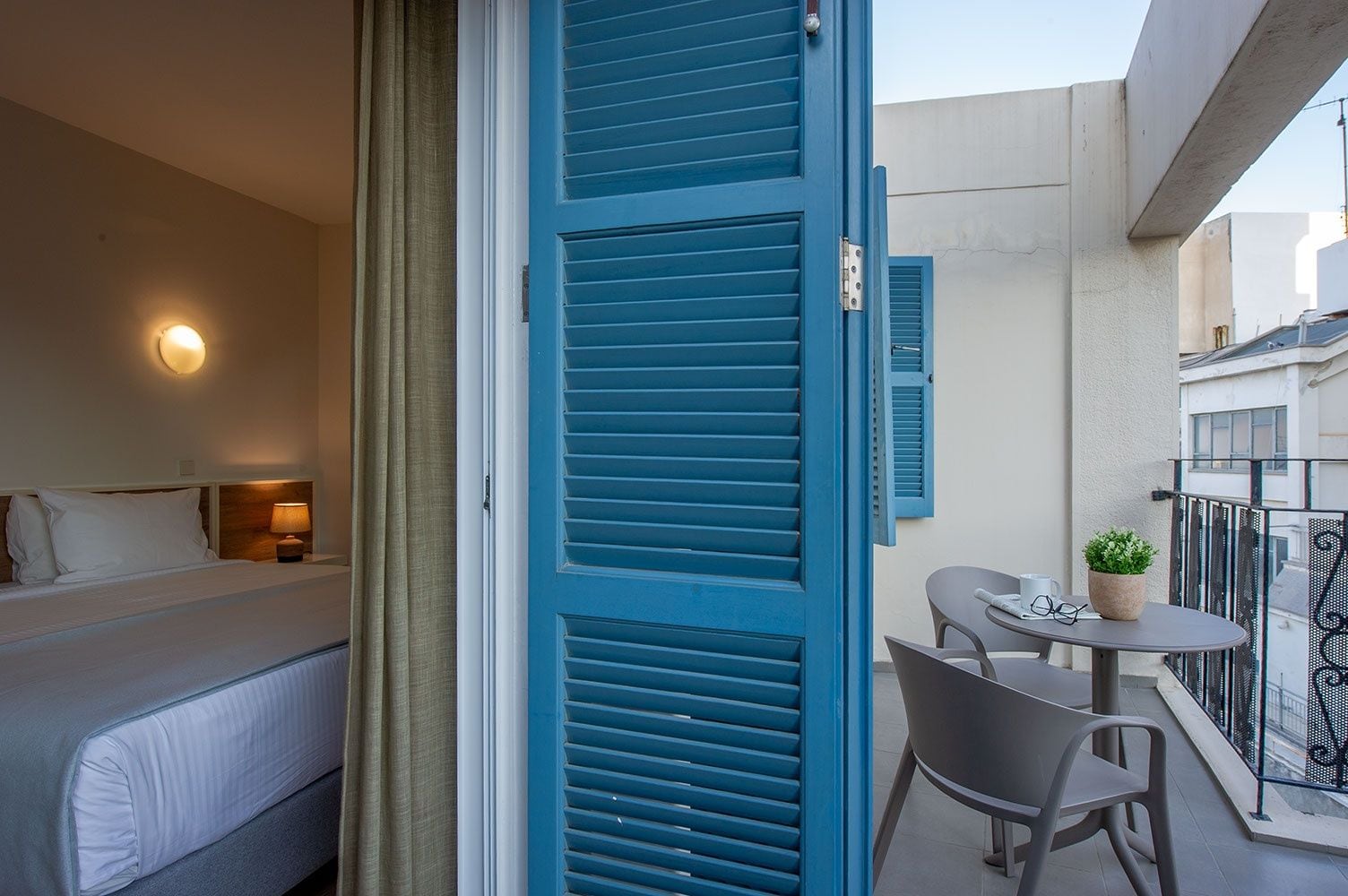 A hotel room with a bed, a nightstand, a lamp, a window with curtains, and a balcony with a table, chairs, and a potted plant.
