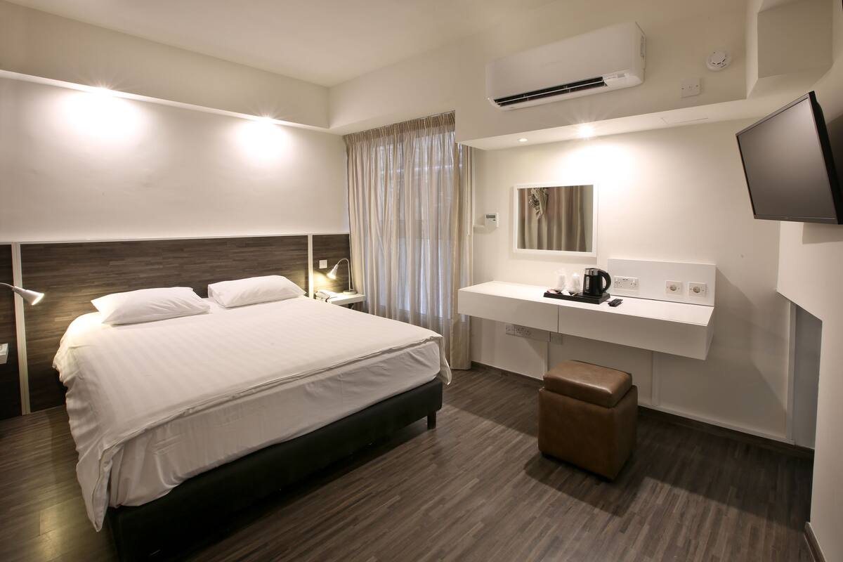 A modern hotel room with a large bed, a bedside table with a lamp, a small seating area, a vanity with a mirror, and a mounted TV.