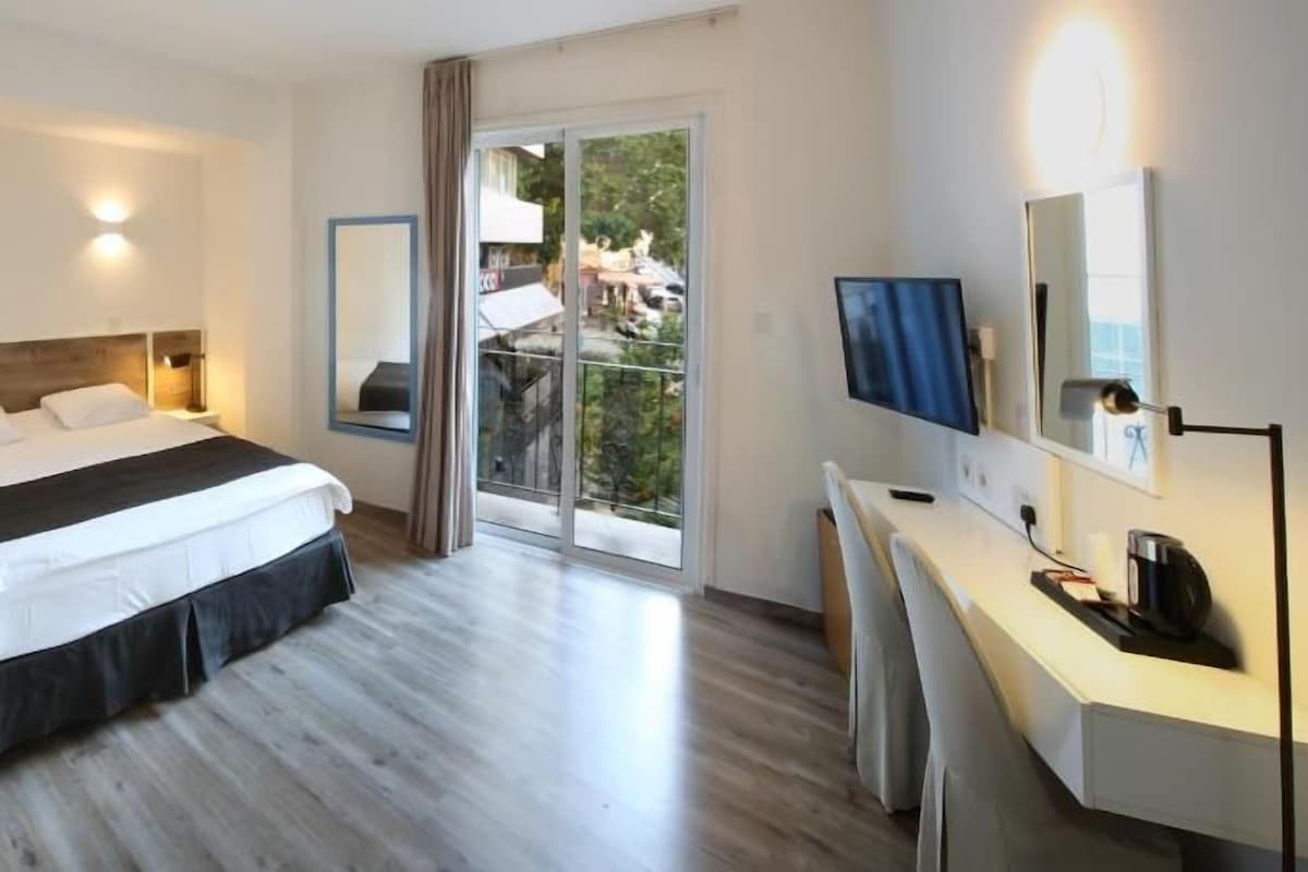 Hotellihuone, jossa on sänky, yöpöydät, työpöytä tuolilla, televisio ja parveke, jolta on näkymä.