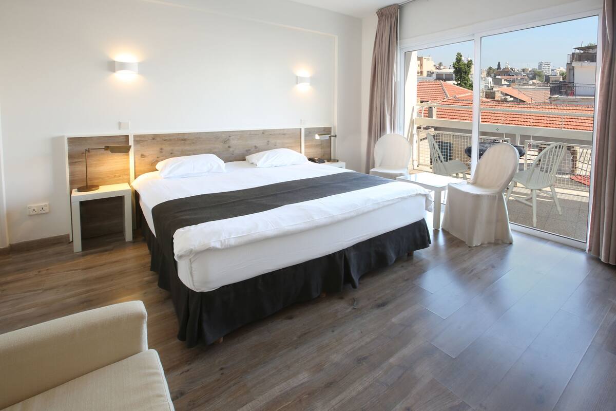 A modern hotel room with a large bed, bedside tables, a chair, and a balcony with a city view.