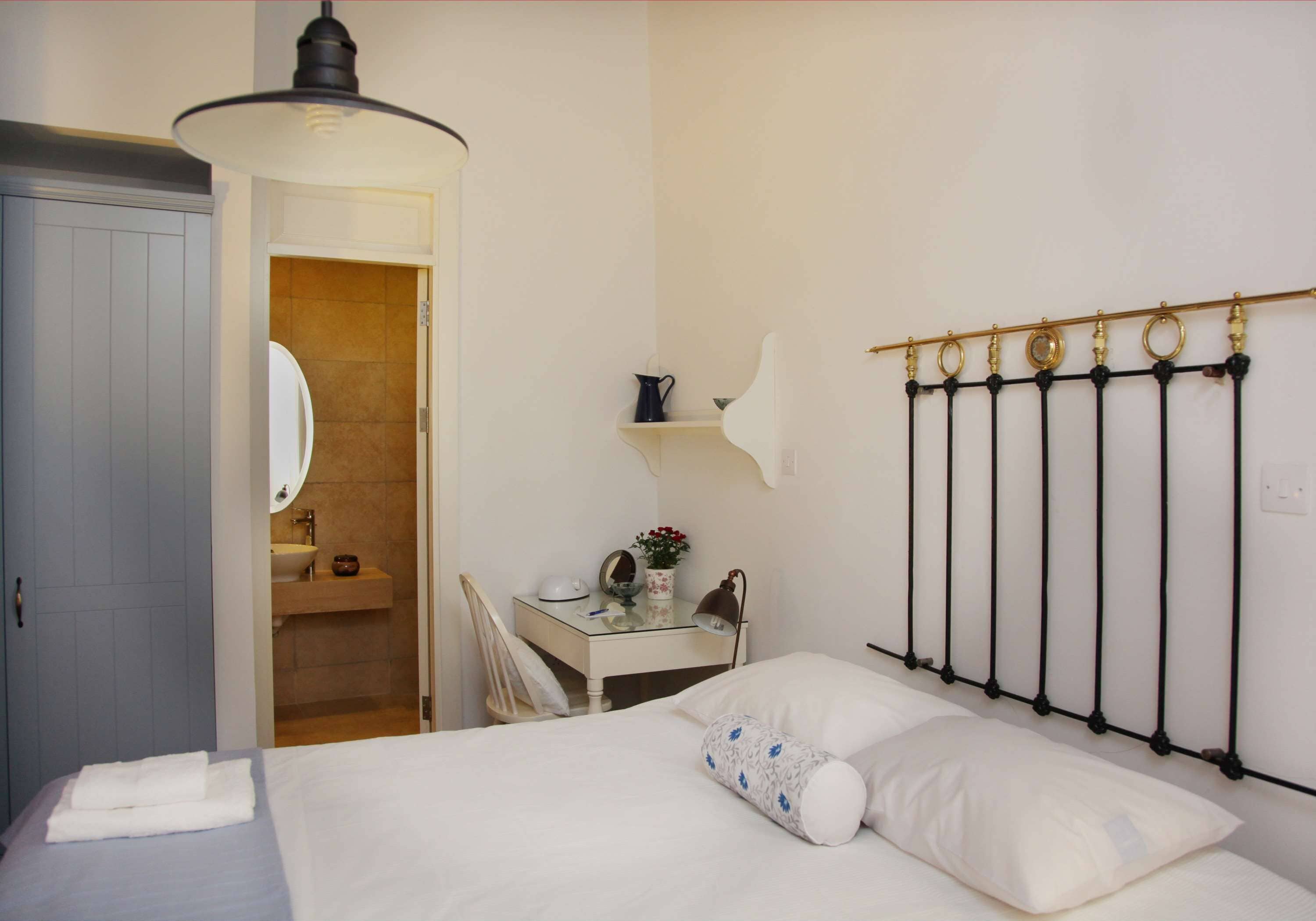 A bedroom with a bed, a desk with a chair, a mirror, a shelf with a vase of flowers, and a radiator.