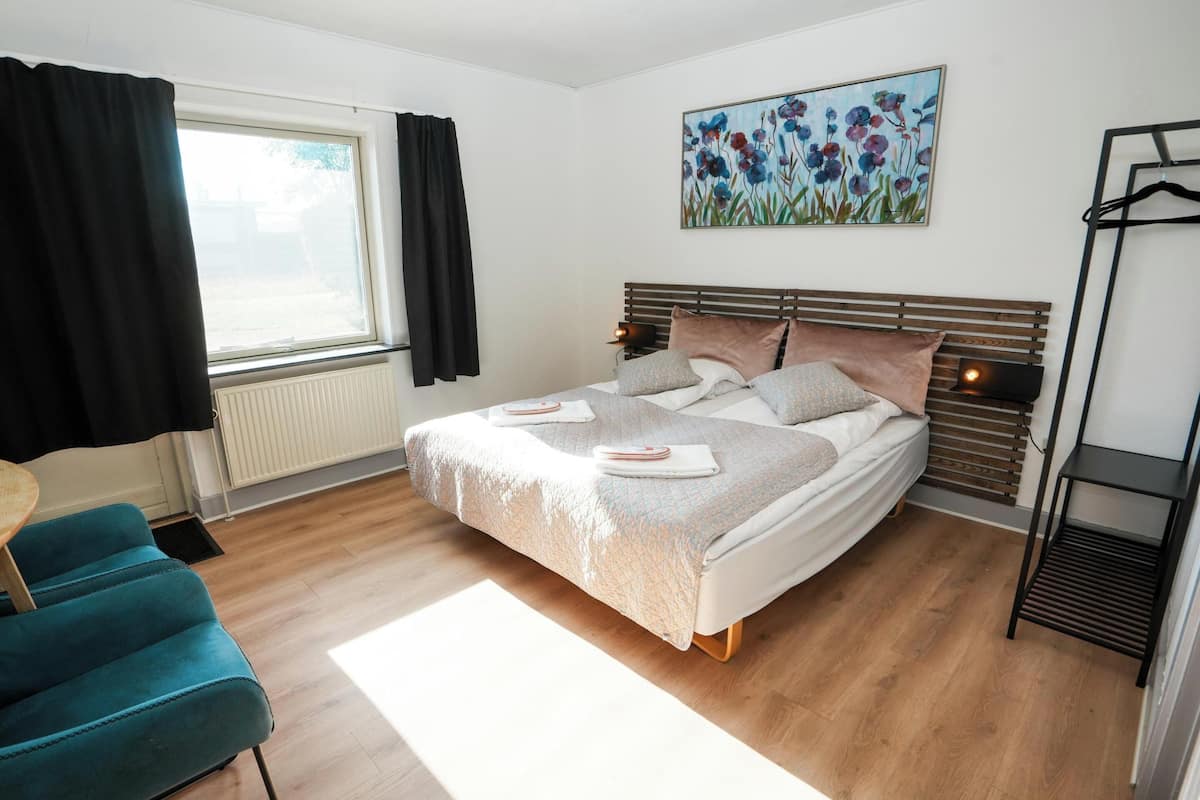 Un dormitorio moderno con una cama de madera, una silla azul, un radiador, una ventana con cortinas negras y un cuadro en la pared.