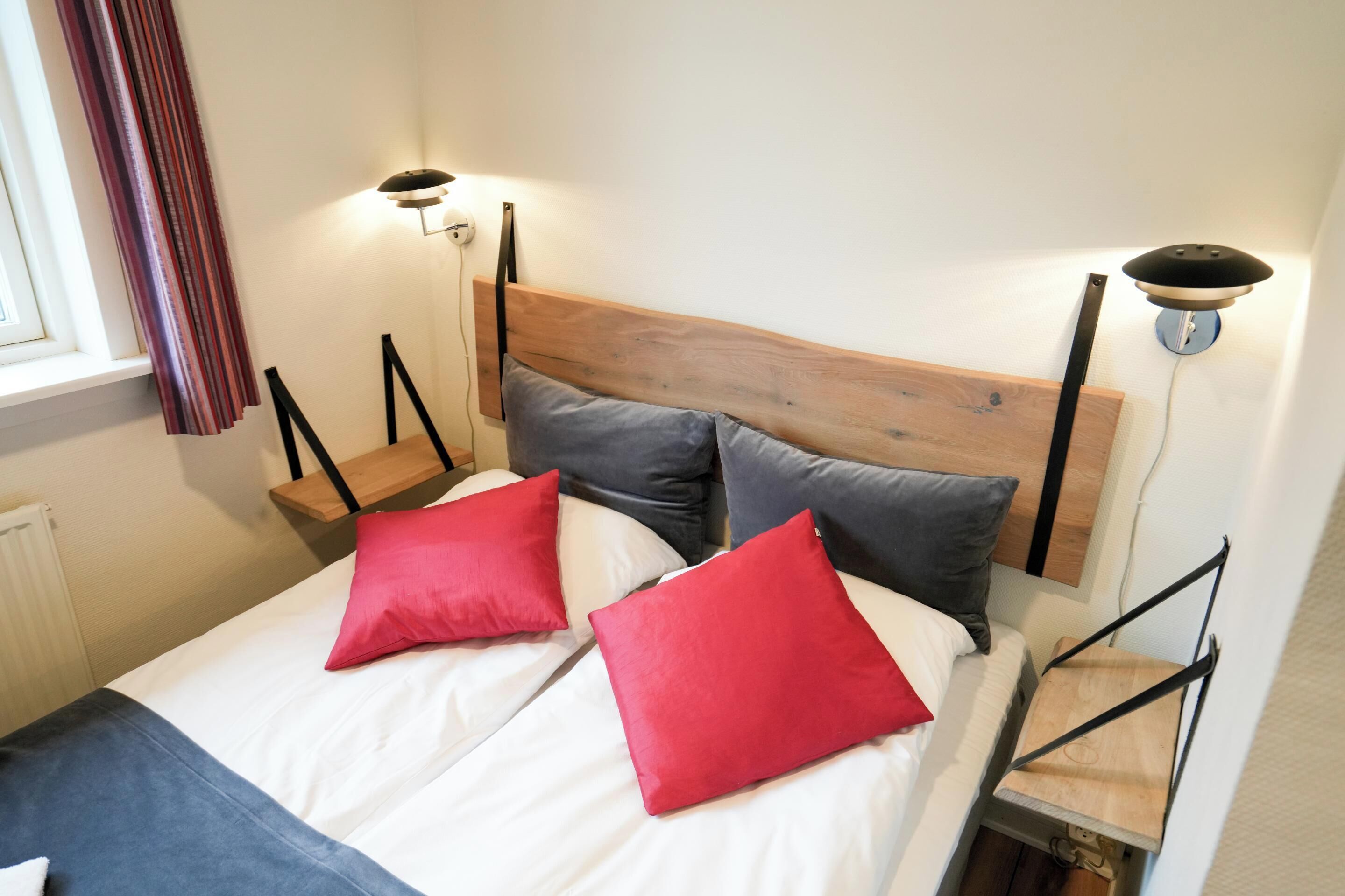Una cama con cabecera de madera, dos almohadas rojas y una almohada gris. Hay dos mesitas de noche fijadas a la pared con lámparas.