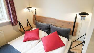 Una cama con cabecera de madera, dos almohadas rojas y una almohada gris. Hay dos mesitas de noche fijadas a la pared con lámparas.