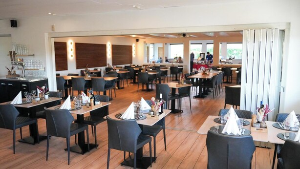Un restaurante espacioso con numerosas mesas y sillas, una zona de bar y grandes ventanales que dejan entrar luz natural.
