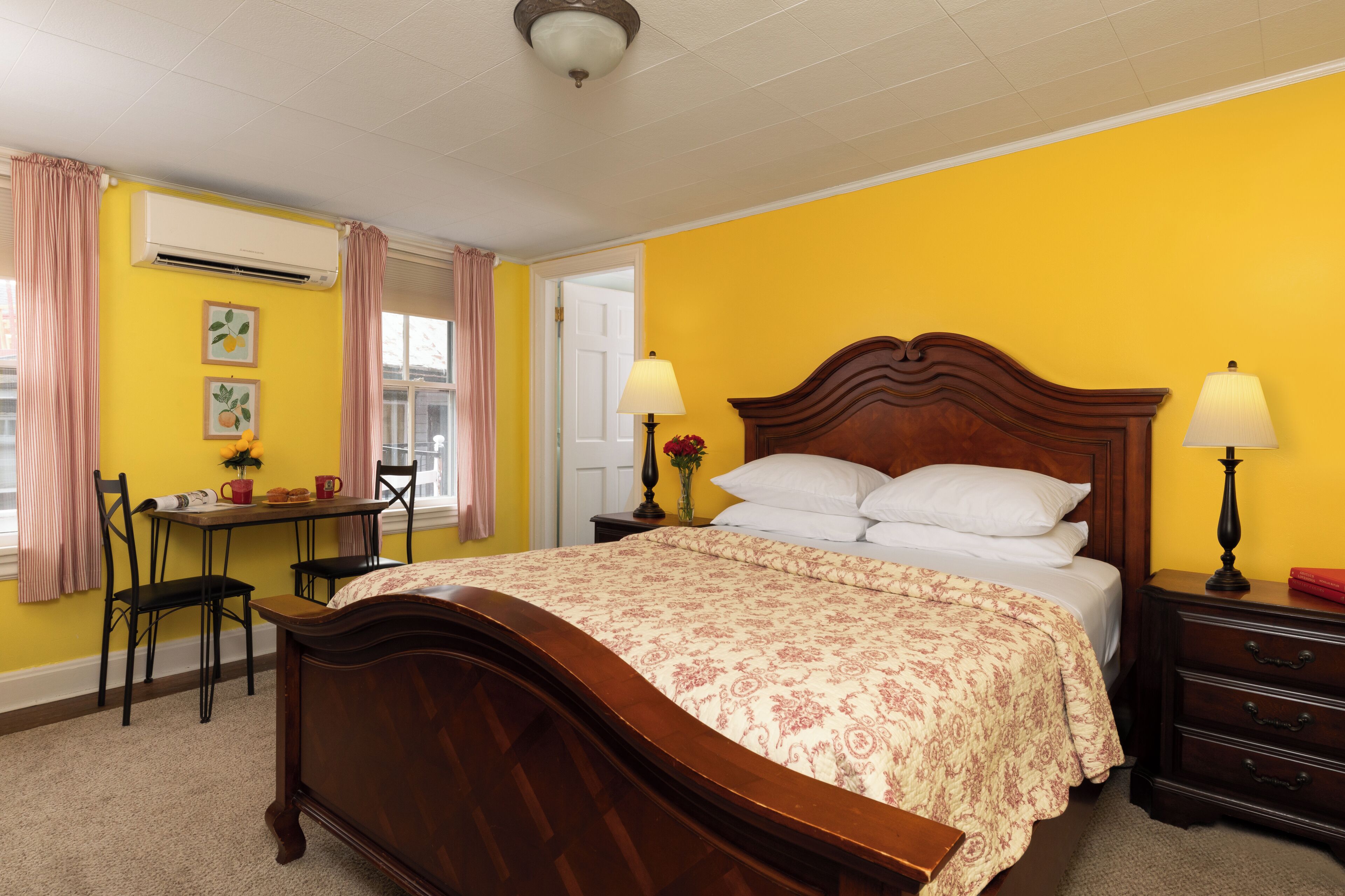A bedroom with a wooden bed, a bedside table with lamps, a chair, a desk, and a window with curtains.