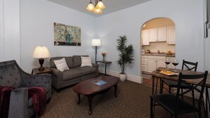 A living room with a sofa, coffee table, and lamp. There is a dining table with chairs and a potted plant. A kitchen is visible through an archway. - Carlisle House (Carlisle)