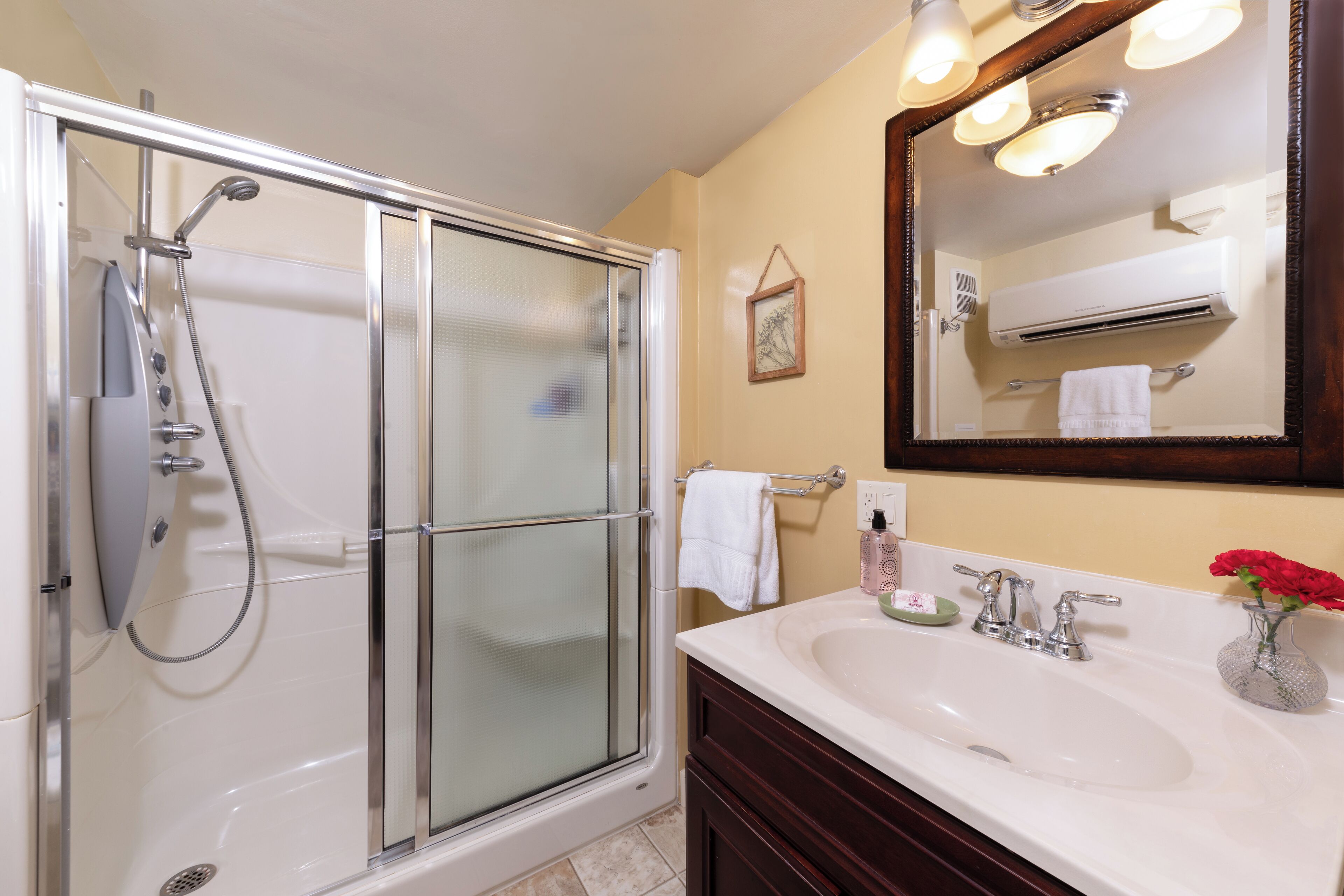 A bathroom with a glass-enclosed shower, a white sink with a vase of flowers, and a mirror with lights above it.