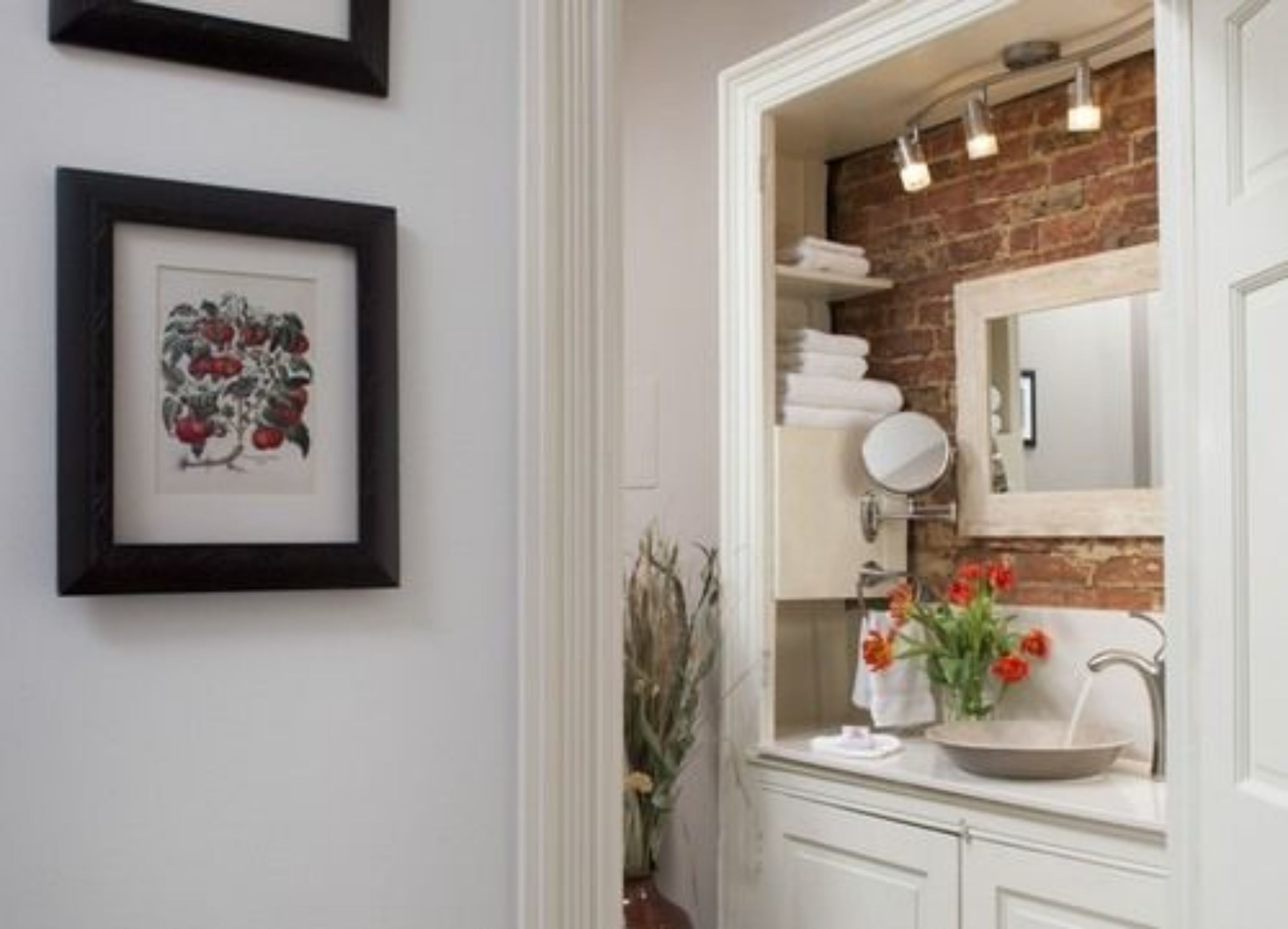 A bathroom with a white vanity, a mirror, a sink, and a brick wall. There are framed pictures on the wall and a vase with flowers on the counter.