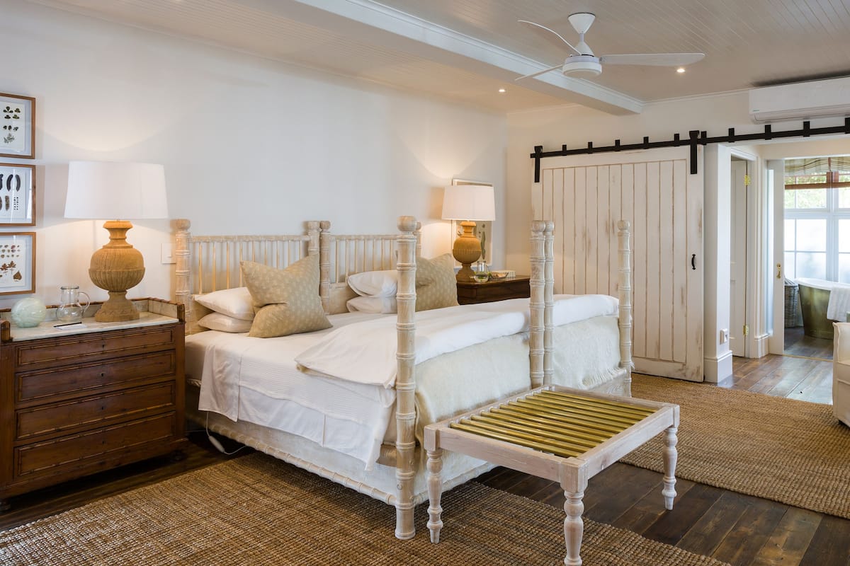 Une chambre à coucher avec un lit à baldaquin, une table de chevet en bois, une petite table de chevet avec une lampe, une chaise et une grande fenêtre donnant sur l’extérieur.
