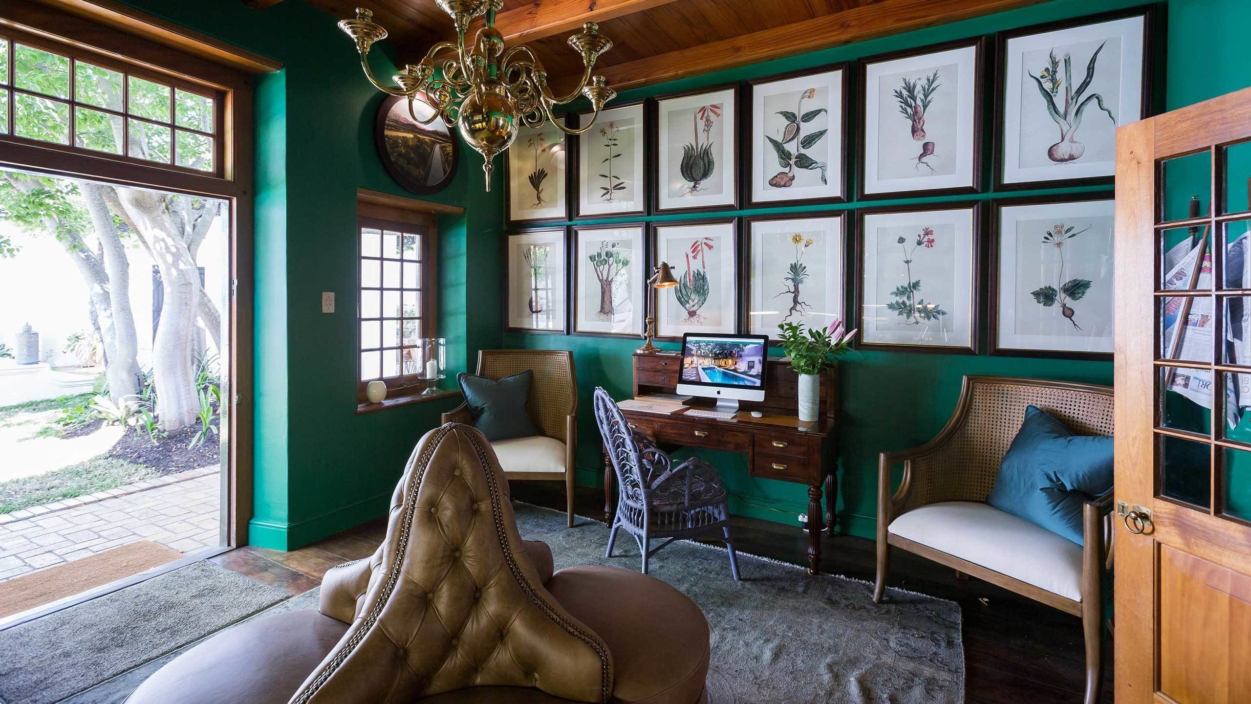 A room with a large window, a desk with a computer, a chair, a sofa, and a wall with framed botanical illustrations.