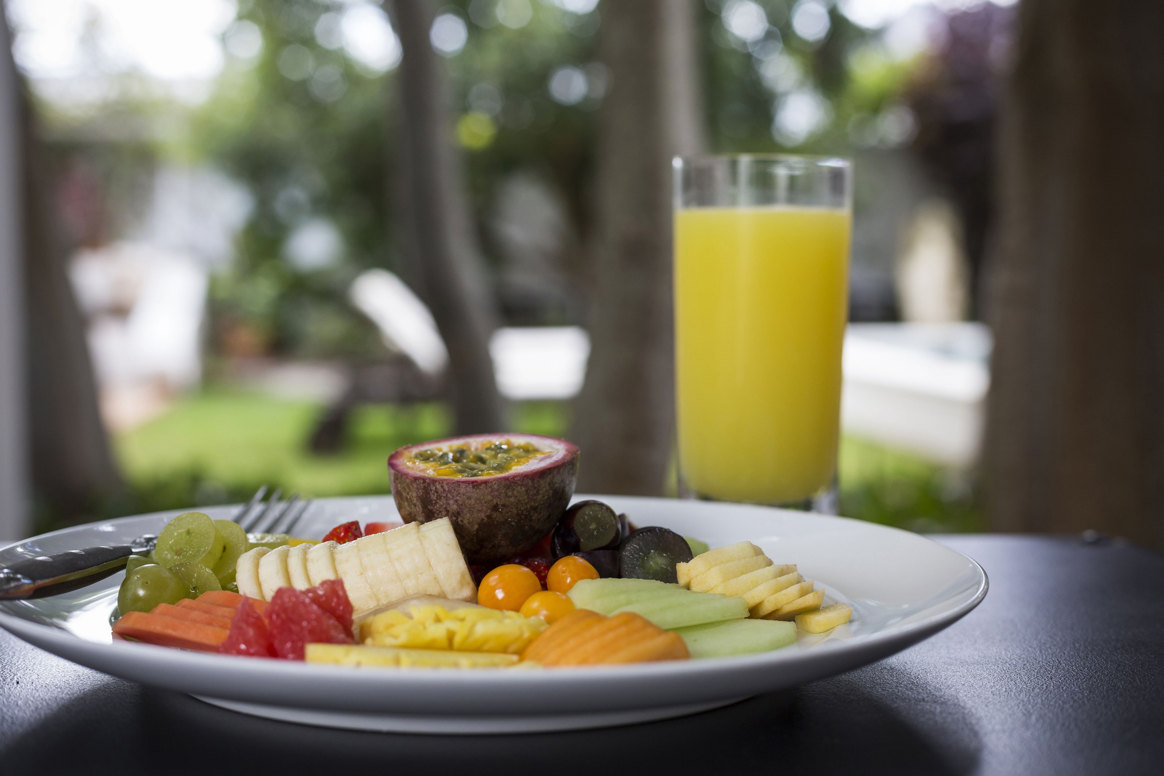 A plate of fruit and a glass of orange juice on a table with a blurred outdoor background.