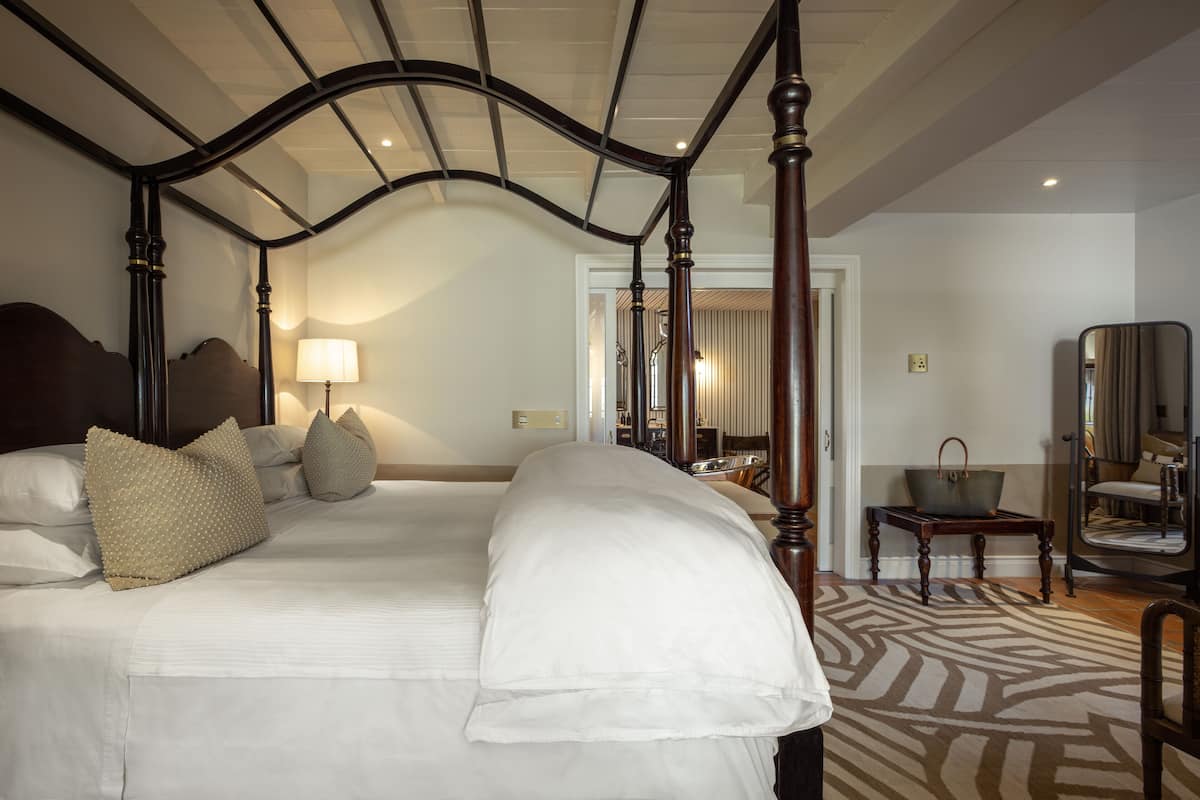Un lit à baldaquin avec du linge de lit blanc et des coussins décoratifs, une tête de lit en bois et un tapis à motifs sur le sol.