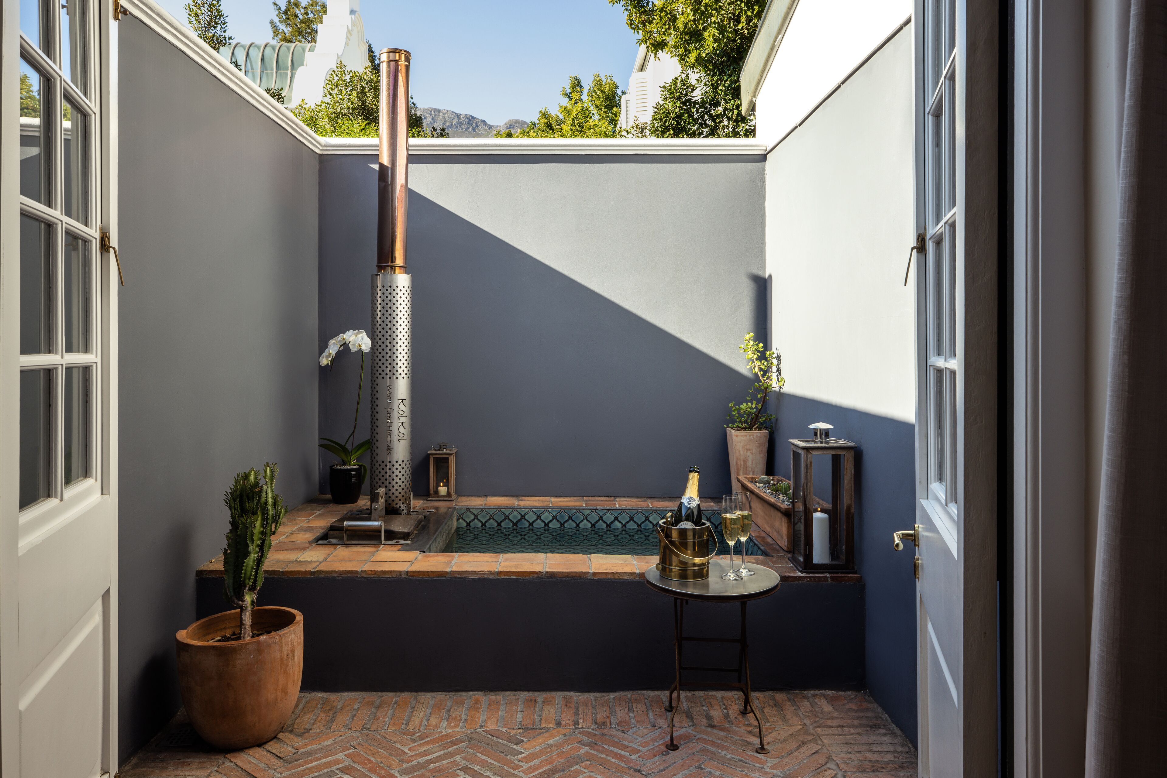 A small, enclosed outdoor space with a brick floor, a small pool, a tall, cylindrical structure, and a table with a bottle and glass.