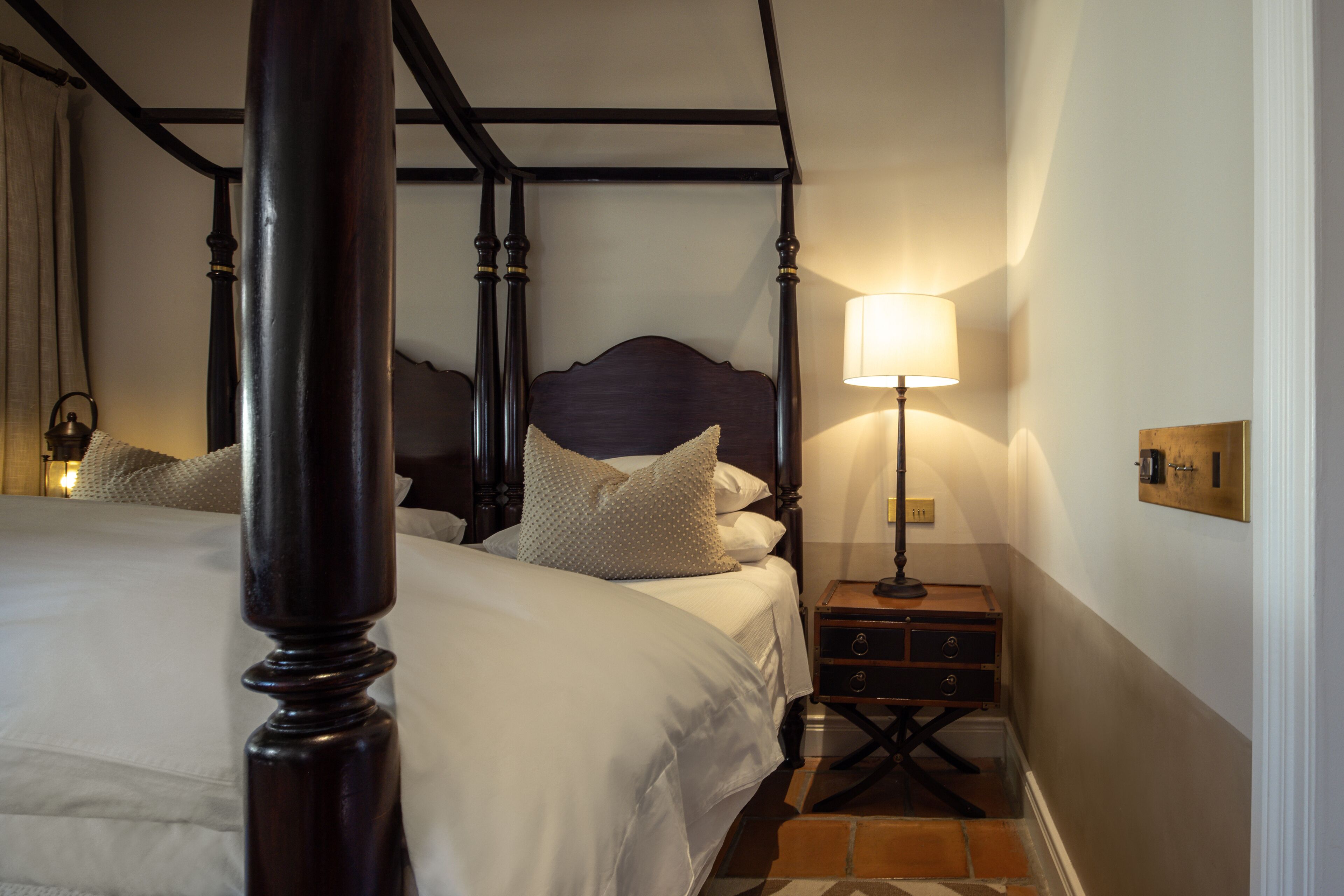 A four-poster bed with a wooden headboard, a bedside table with a lamp, and a nightstand with drawers.
