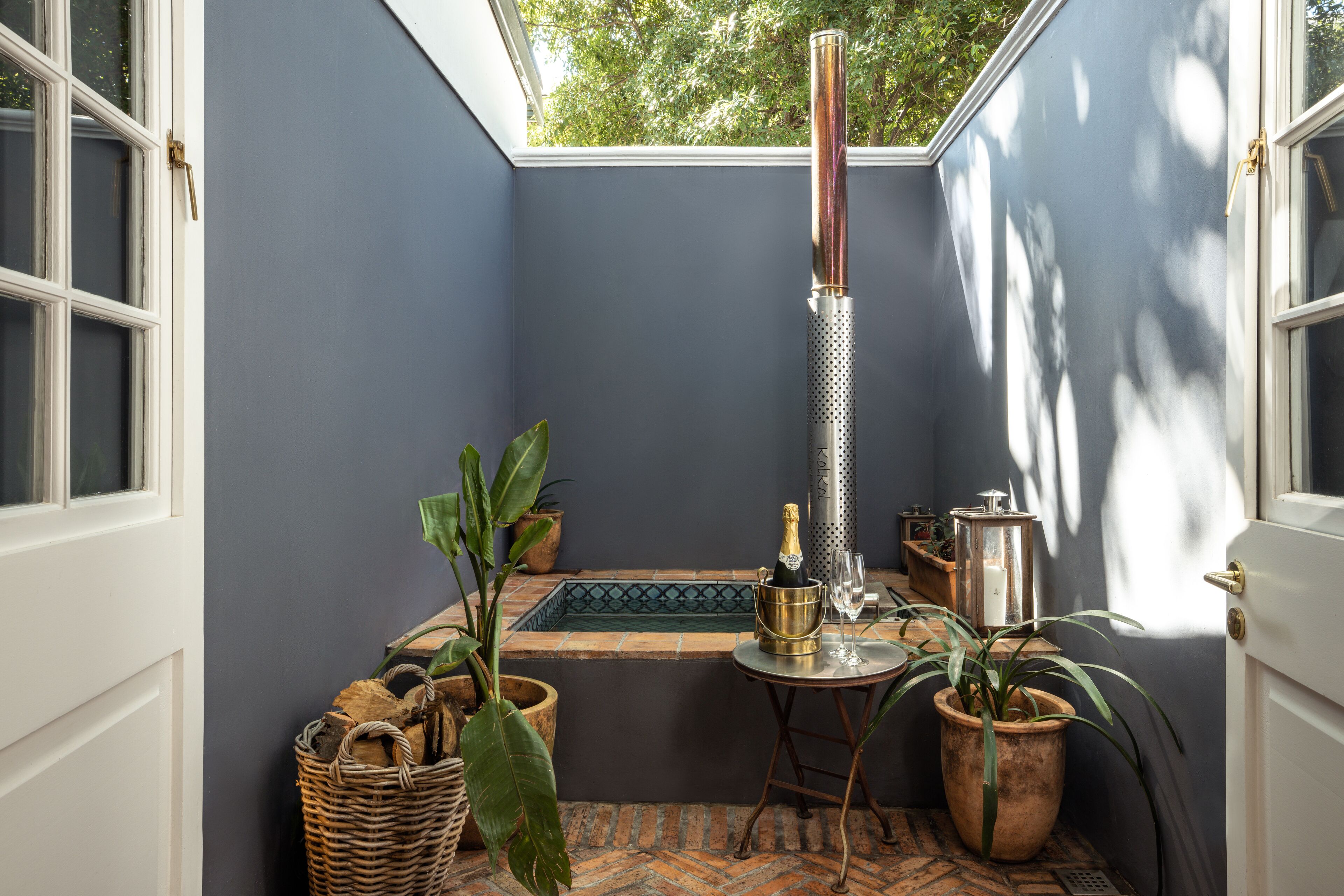 A corner room with a bathtub, a small table with a bottle and glass, and potted plants.