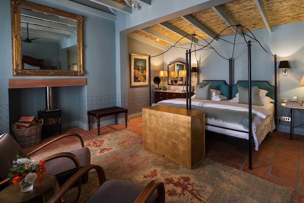 Une chambre avec un grand lit, un banc en bois, une chaise, un miroir, une cheminée et un plafond avec des poutres apparentes.