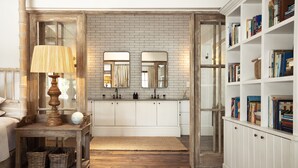 Une pièce avec un plancher en bois, une armoire blanche remplie de livres, une table en bois avec une lampe, et un miroir au-dessus de l’armoire.