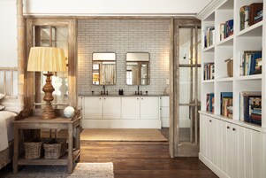 A room with a wooden floor, a white cabinet with books, a wooden table with a lamp, and a mirror above the cabinet.
