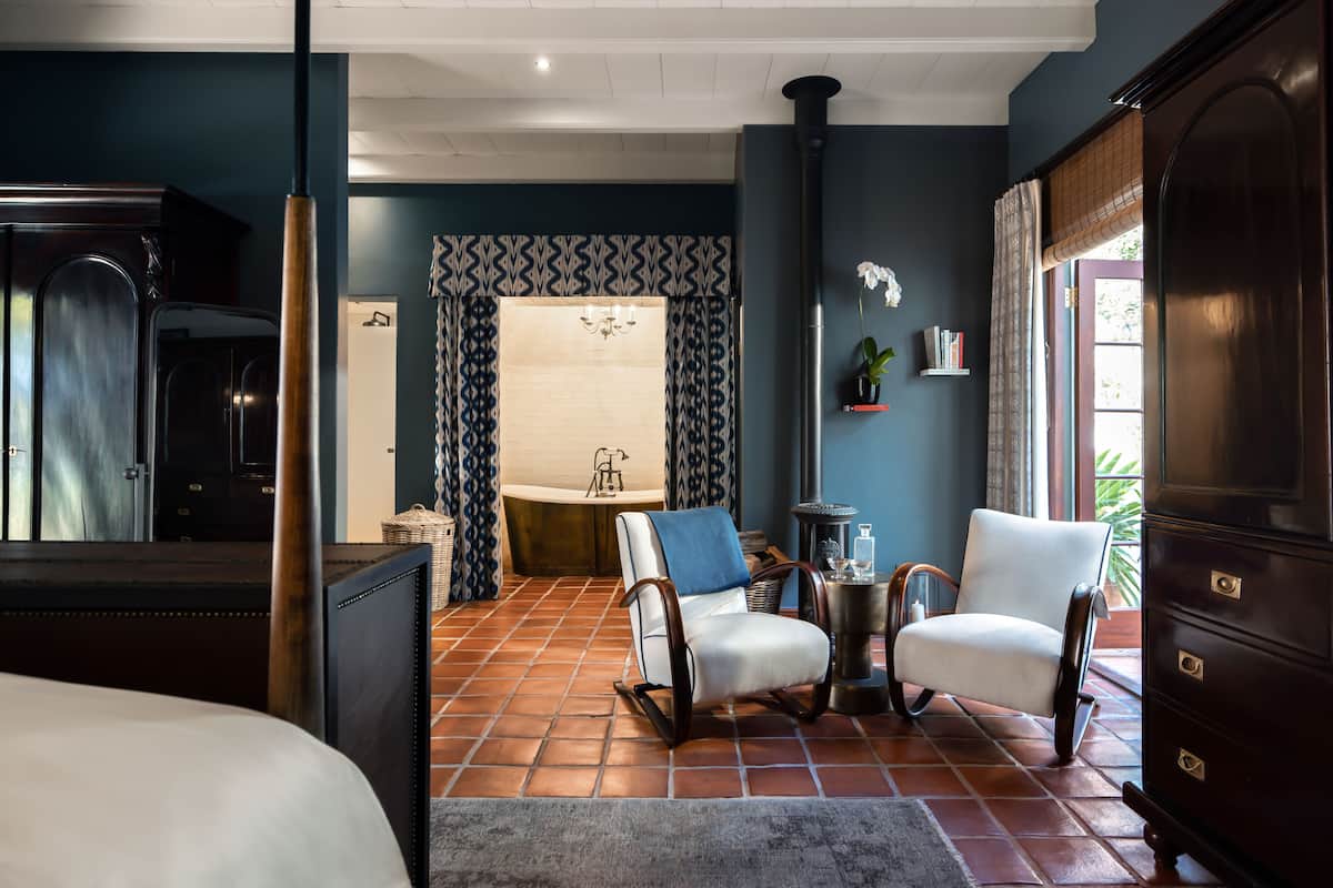 Une pièce comprenant un lit, deux fauteuils, une petite table, une cheminée et une vue sur l’extérieur.