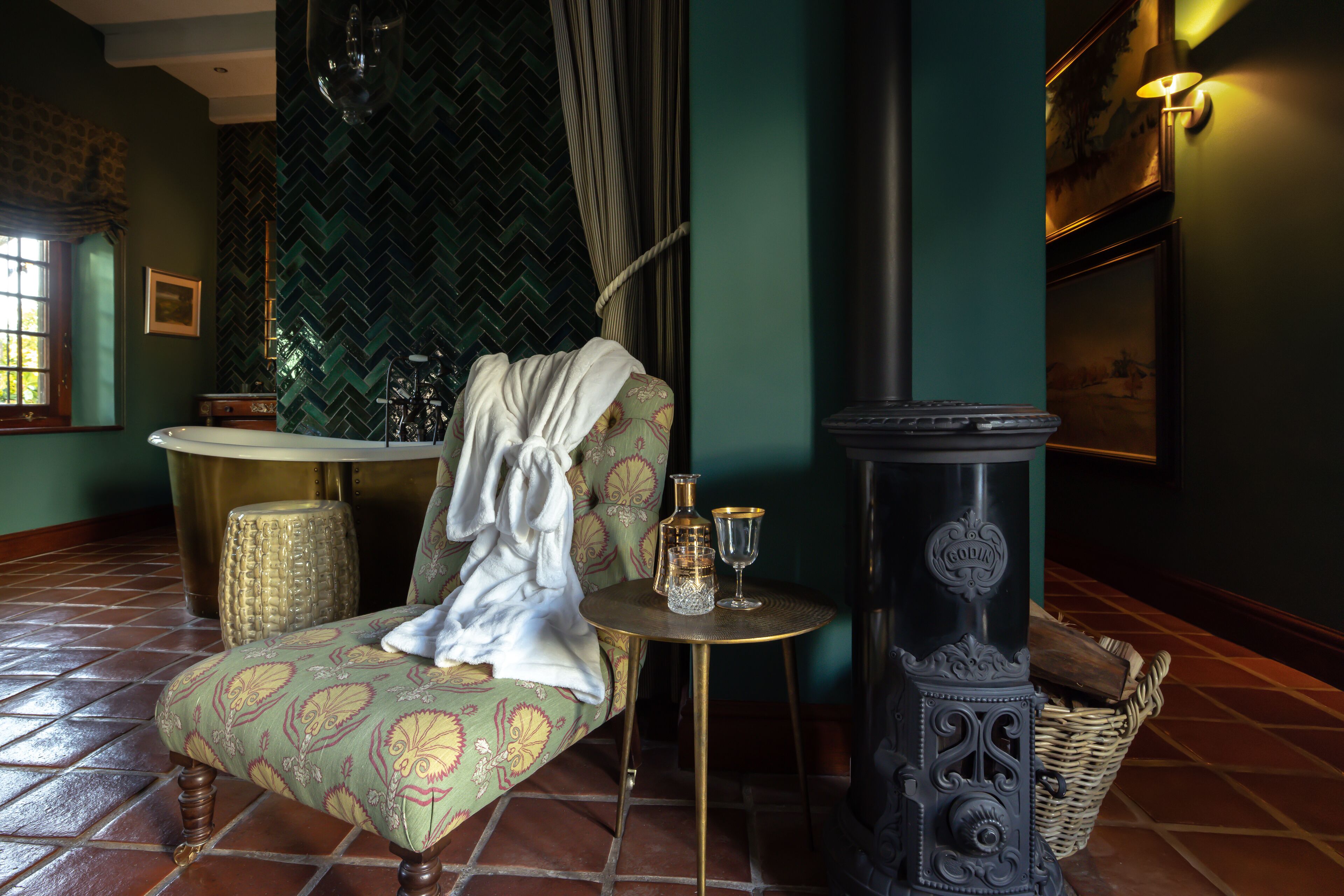 Una habitación con un sillón estampado, una mesita redonda con una botella y un vaso, una estufa decorativa y una cesta de mimbre.