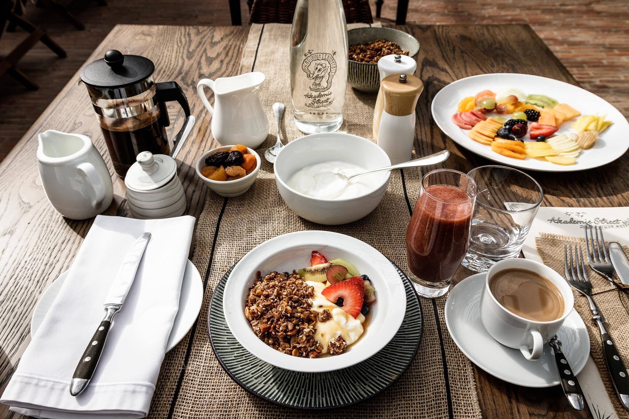 Une table en bois dressée pour le petit-déjeuner, avec une cafetière, un pichet, un bol de yaourt, une assiette de fruits et une tasse à café.