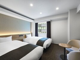 A hotel room with two beds, a small round table, and a view of the city through a window.