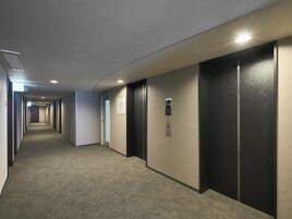 Un pasillo de hotel con pisos alfombrados, una fila de puertas y un ascensor con pantalla digital.
