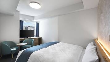 A hotel room with a bed, a desk with a TV, a chair, and a view of the city through the window.