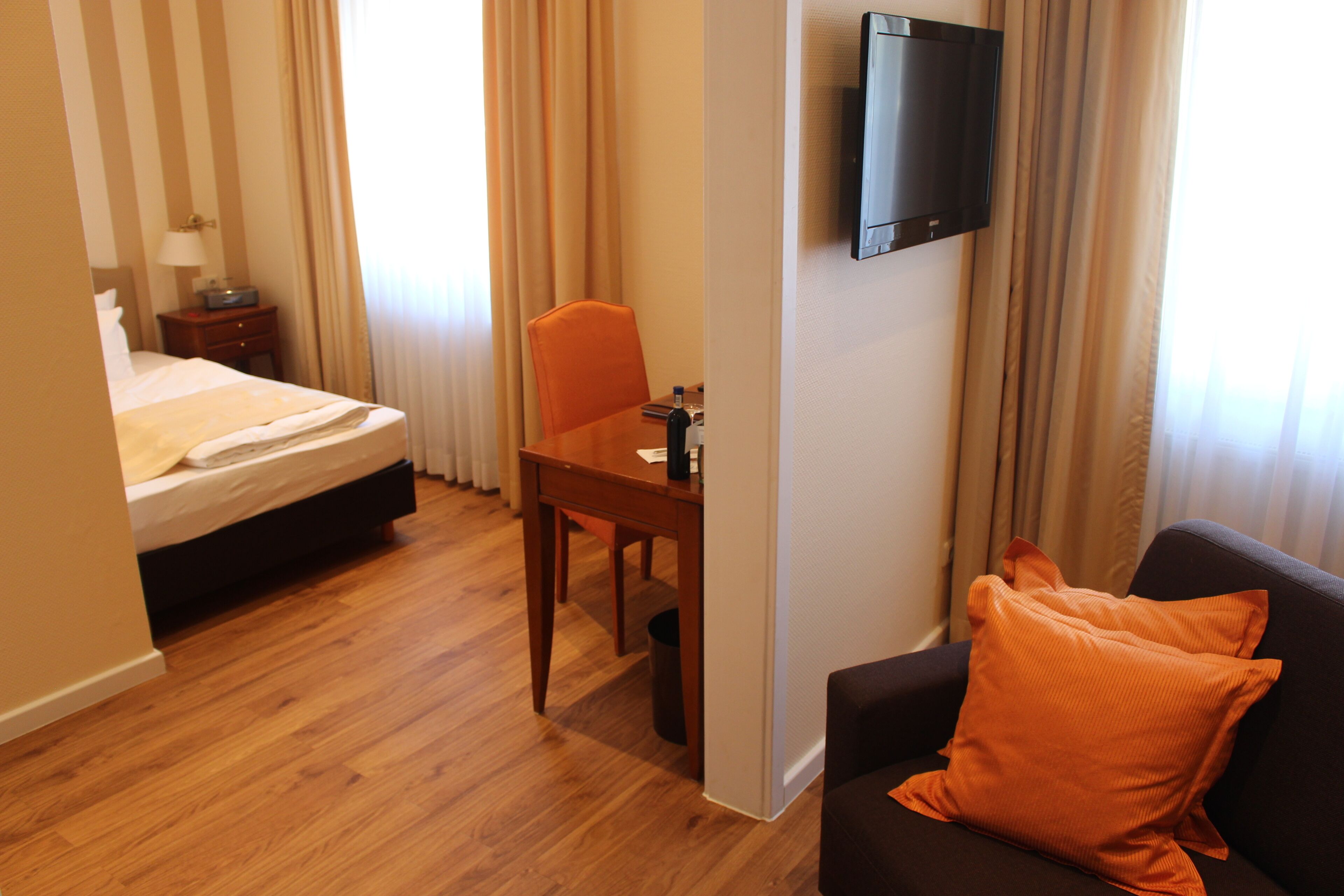 Ein Hotelzimmer mit Bett, Schreibtisch, Stuhl, Fernseher und einer Couch mit orangefarbenem Kissen.