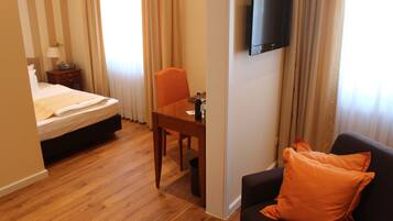 Ein Hotelzimmer mit Bett, Schreibtisch, Stuhl, Fernseher und einer Couch mit orangefarbenem Kissen.
